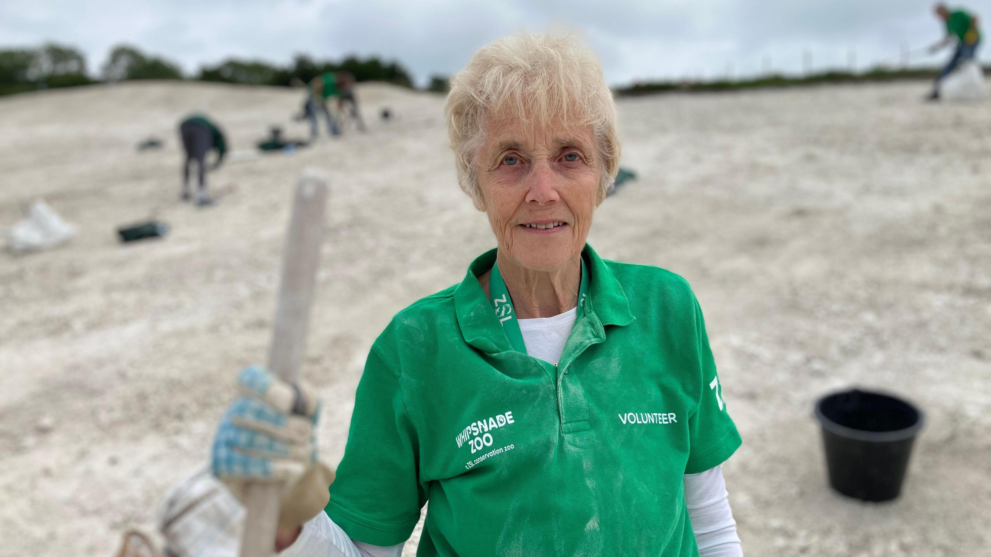 Head and shoulders shot of Anne James, wearing a green t-shirt which says Whipsnade Zoo and volunteer on it. She is holding the handle of a rake. She is smiling at the camera. Behind her, blurred out, is the white chalk ground and people working on it. There is a black bucket behind her in the foreground.