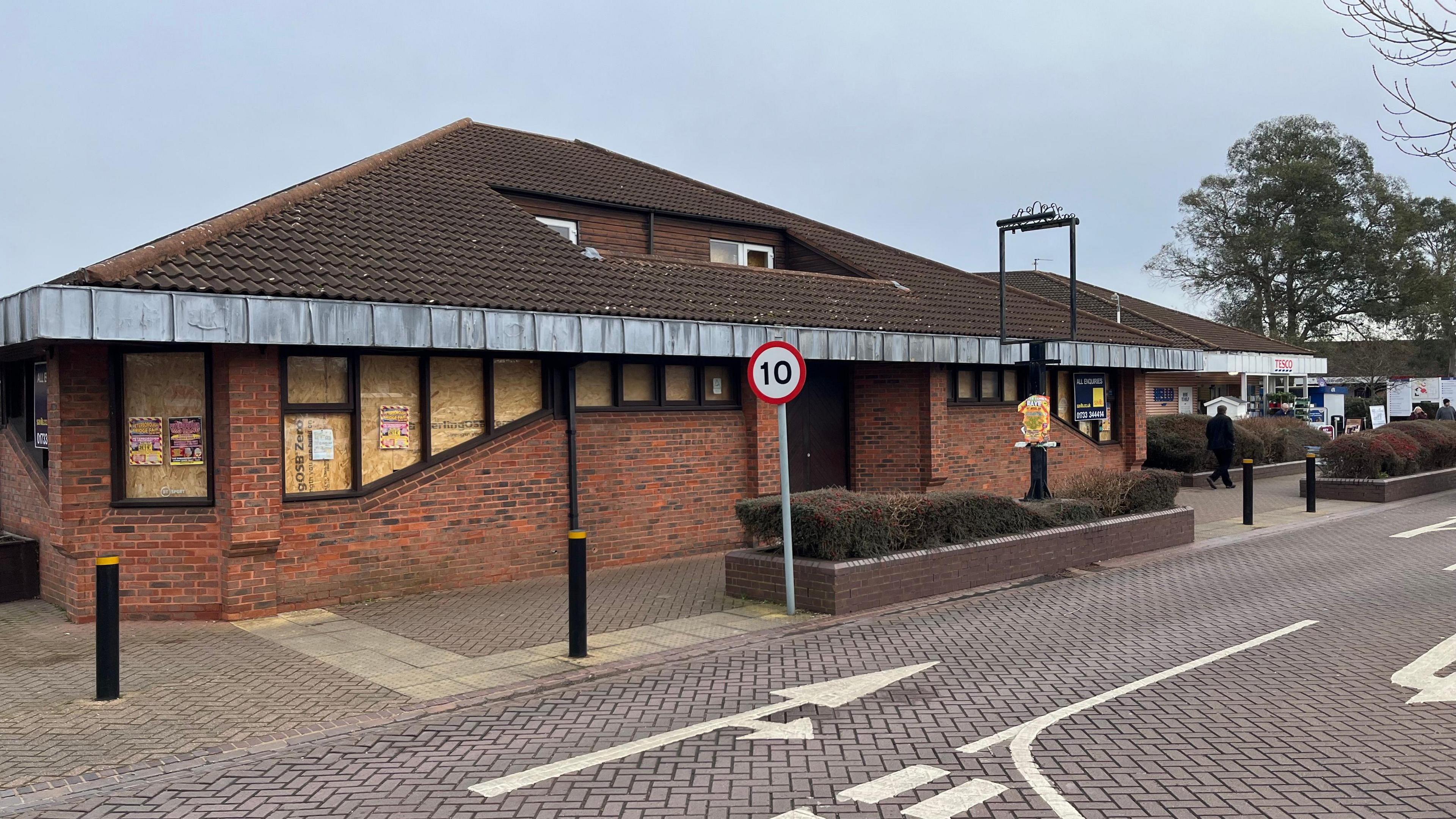 Single story building, opened in 1982 as a pub, it has dark roof tiles and reddish brickwork, there is a metal pub signpost outside, with no sign and road markings on a reddish block paved roadway in the foreground