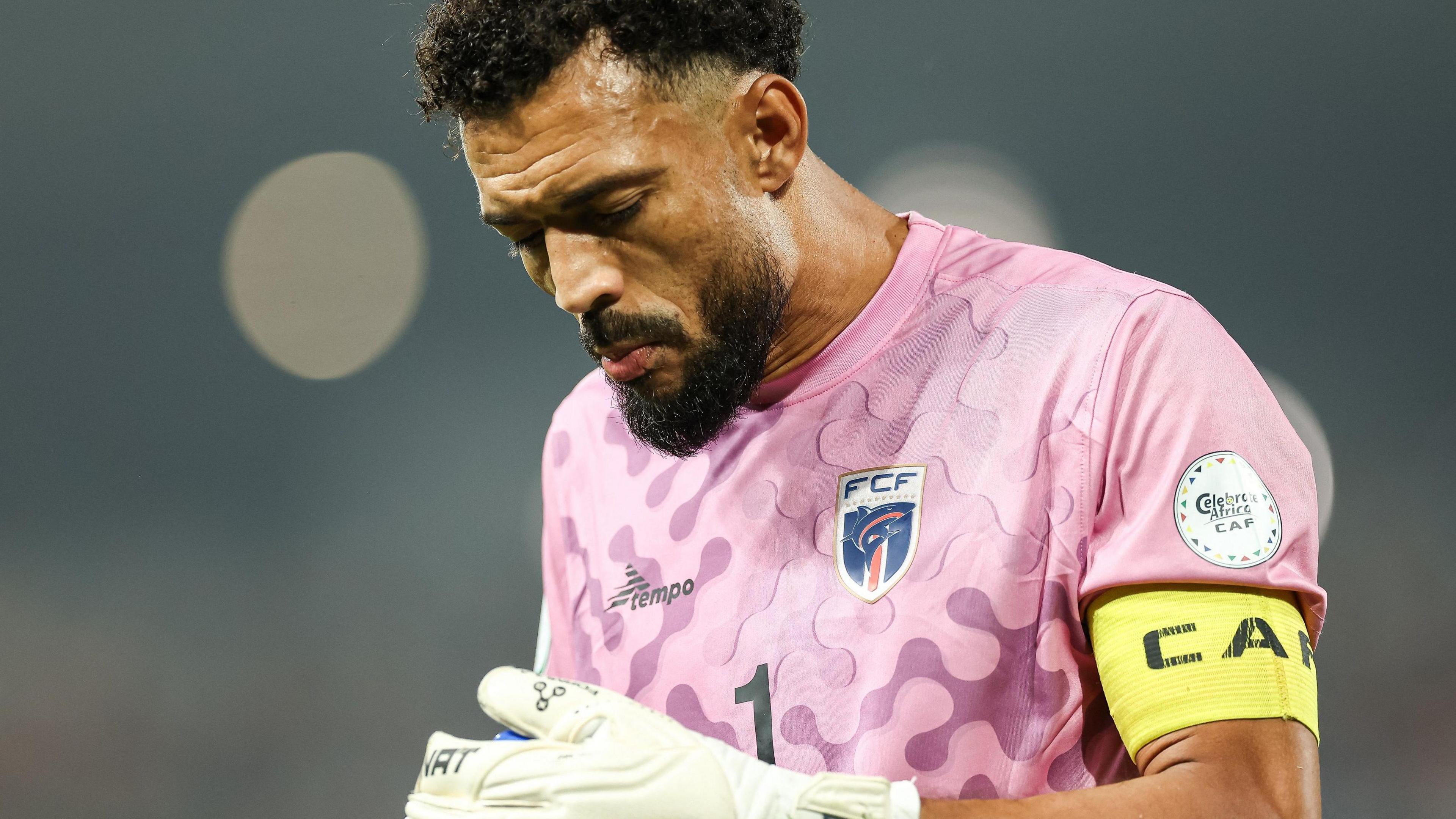 A bearded Cape Verde goalkeeper Vozinha, seen from waist up in a pink national team jersey and yellow armband, looks down at his white gloves