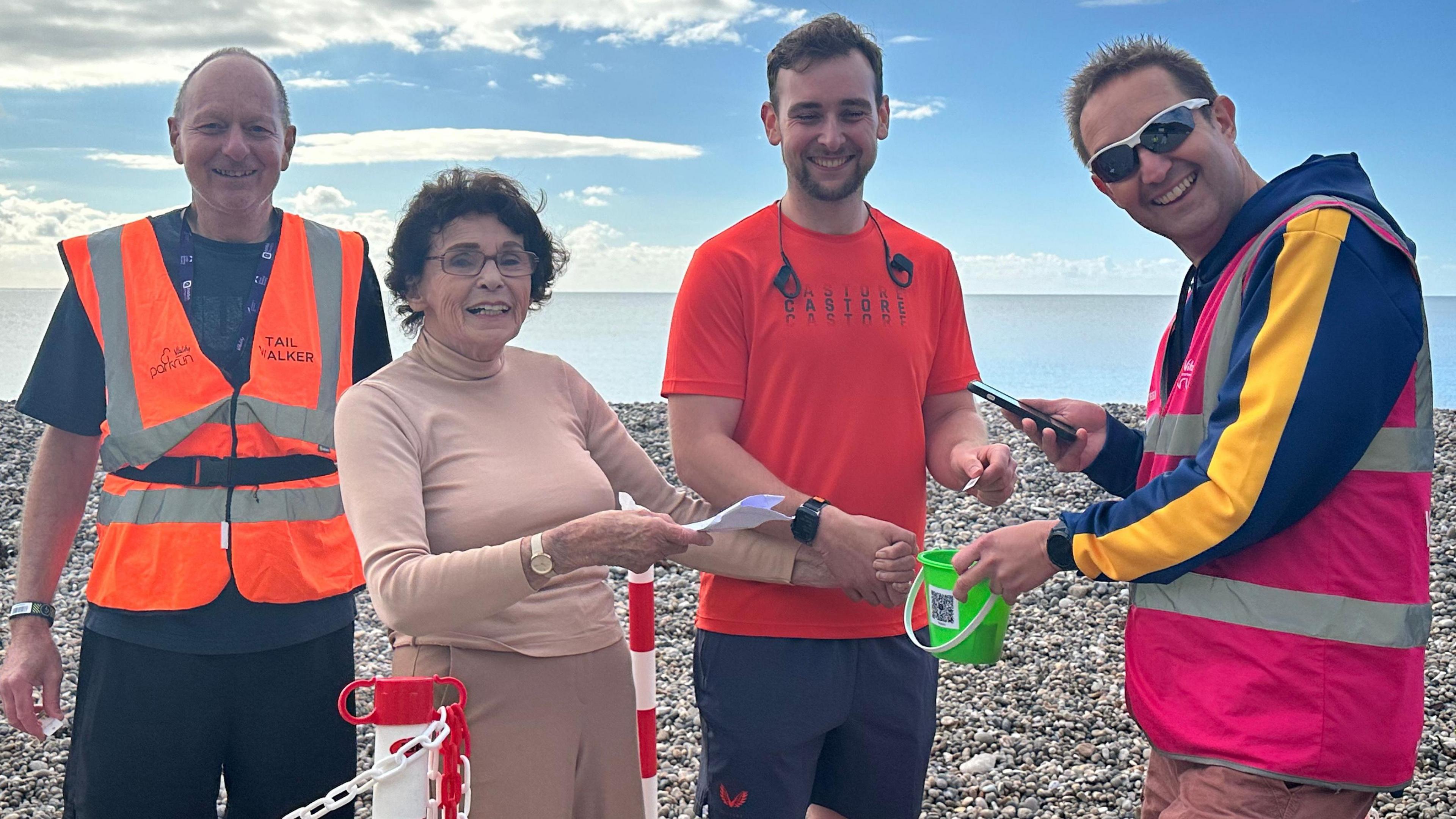 Joan Norcombe at the end of the Seaton Parkrun with three male marshals, two of whom are wearing high-vis jackets.