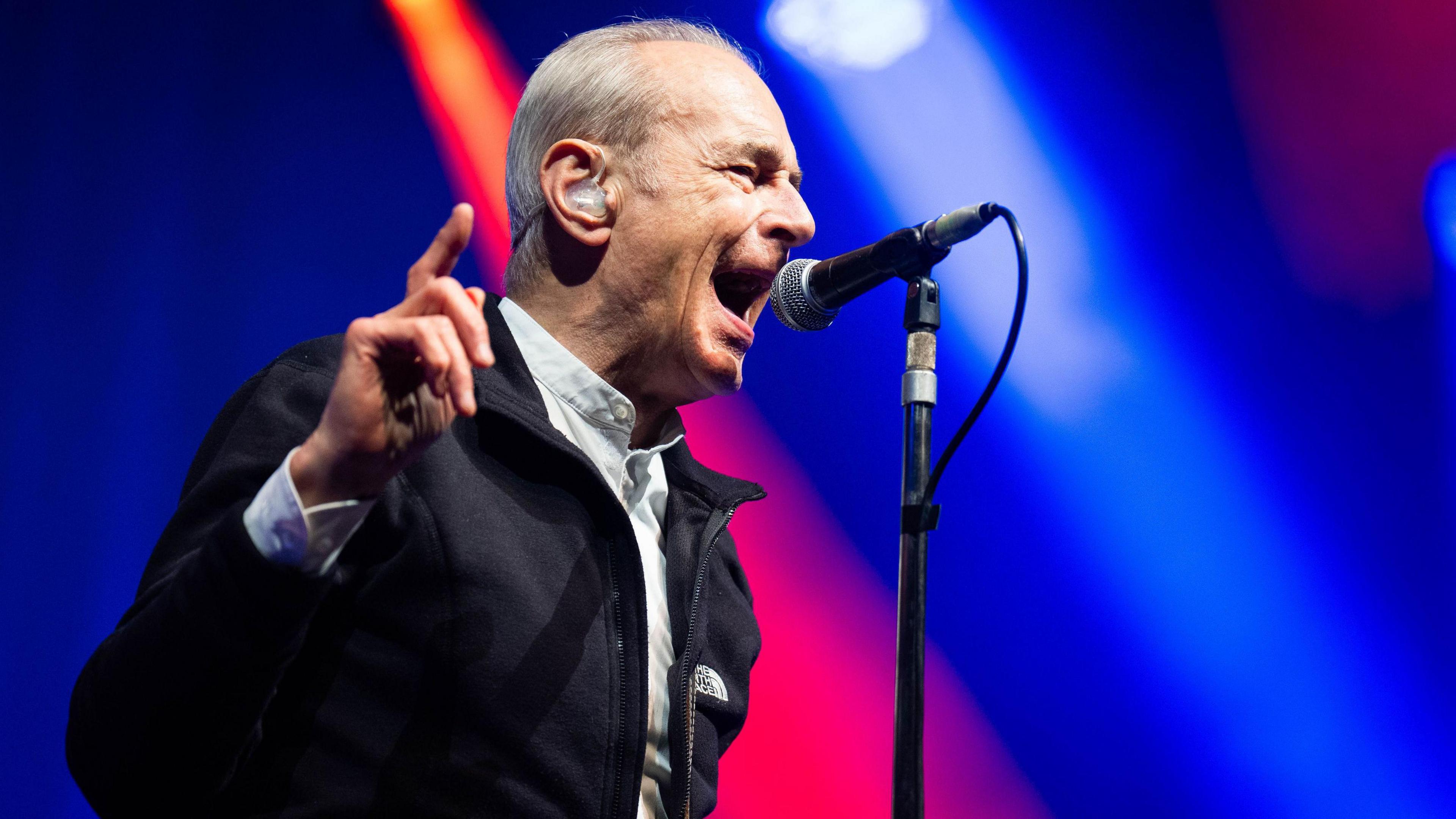 Francis Rossi sings onstage at Vivary Park wearing a dark jacket and pointing a finger in the air, with blue and pink lights behind him