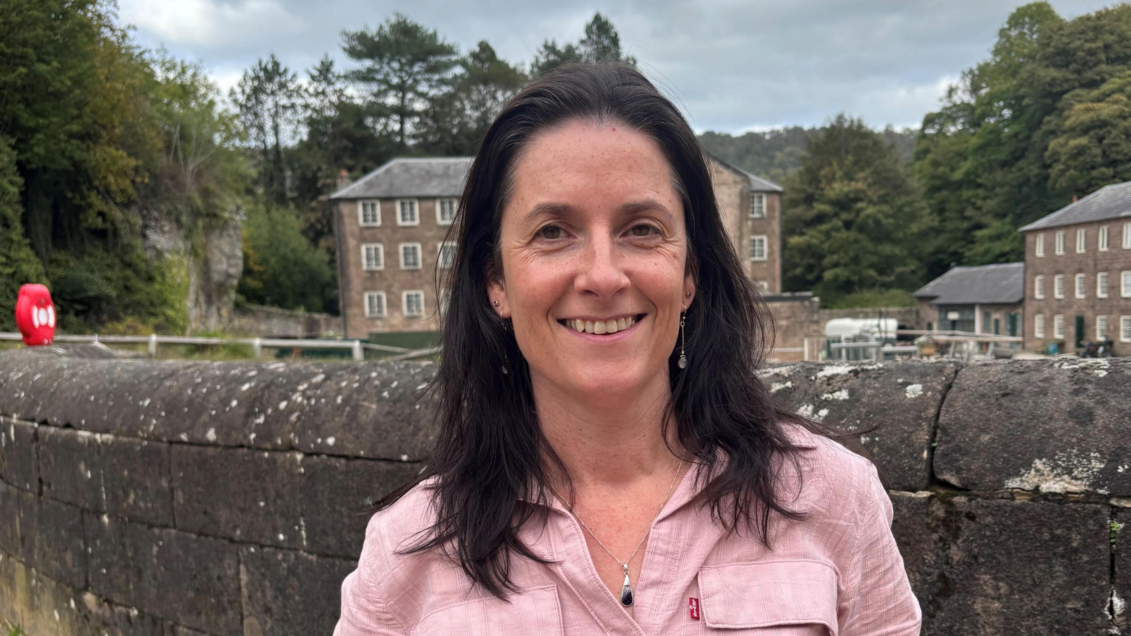 Eilis Scott wearing a pink blouse looking straight at camera in front of walls and buildings of Cromford Mills World Heritage site