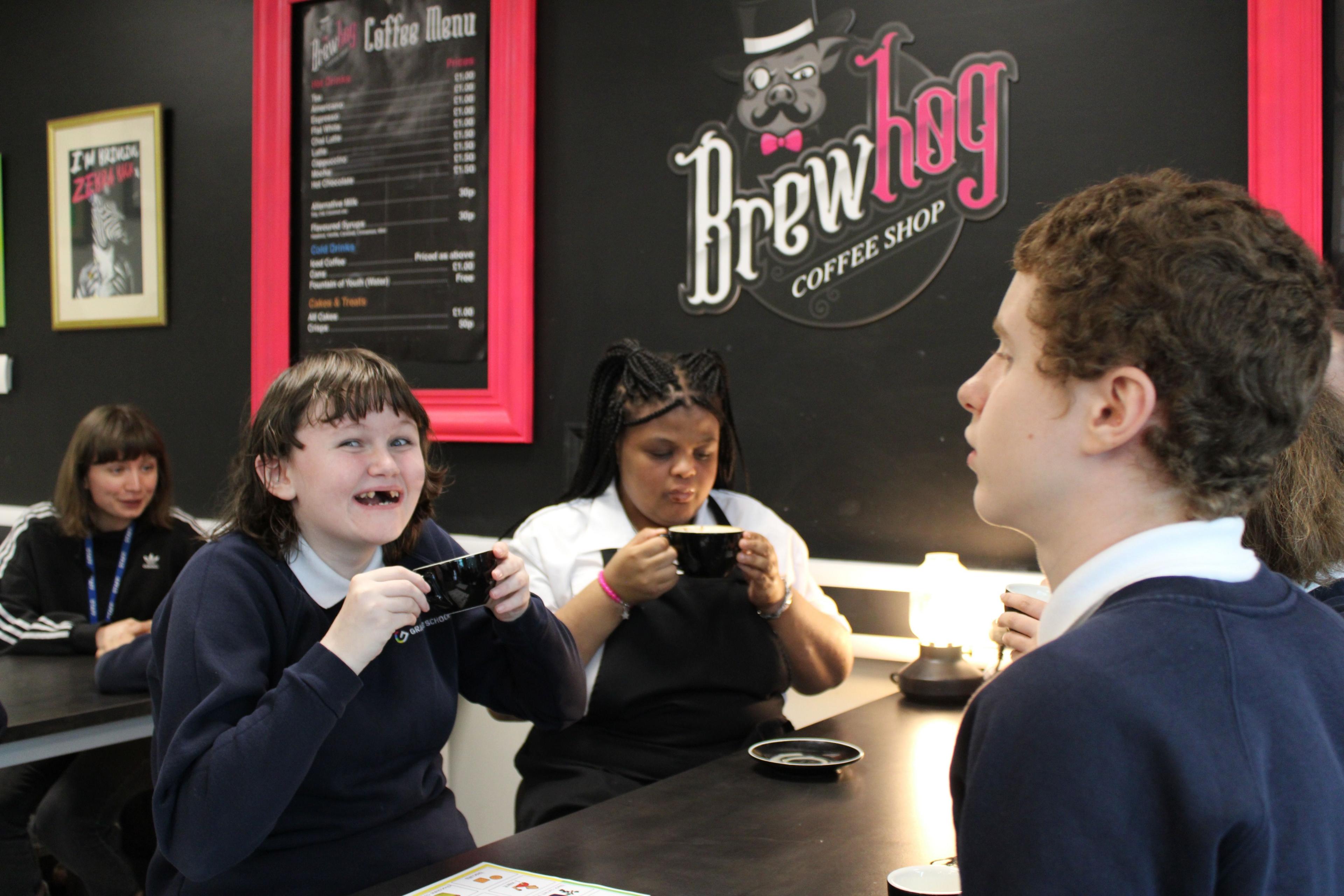 Three pupils at a table in the cafe drinking hot drinks. One is smiling at the camera.