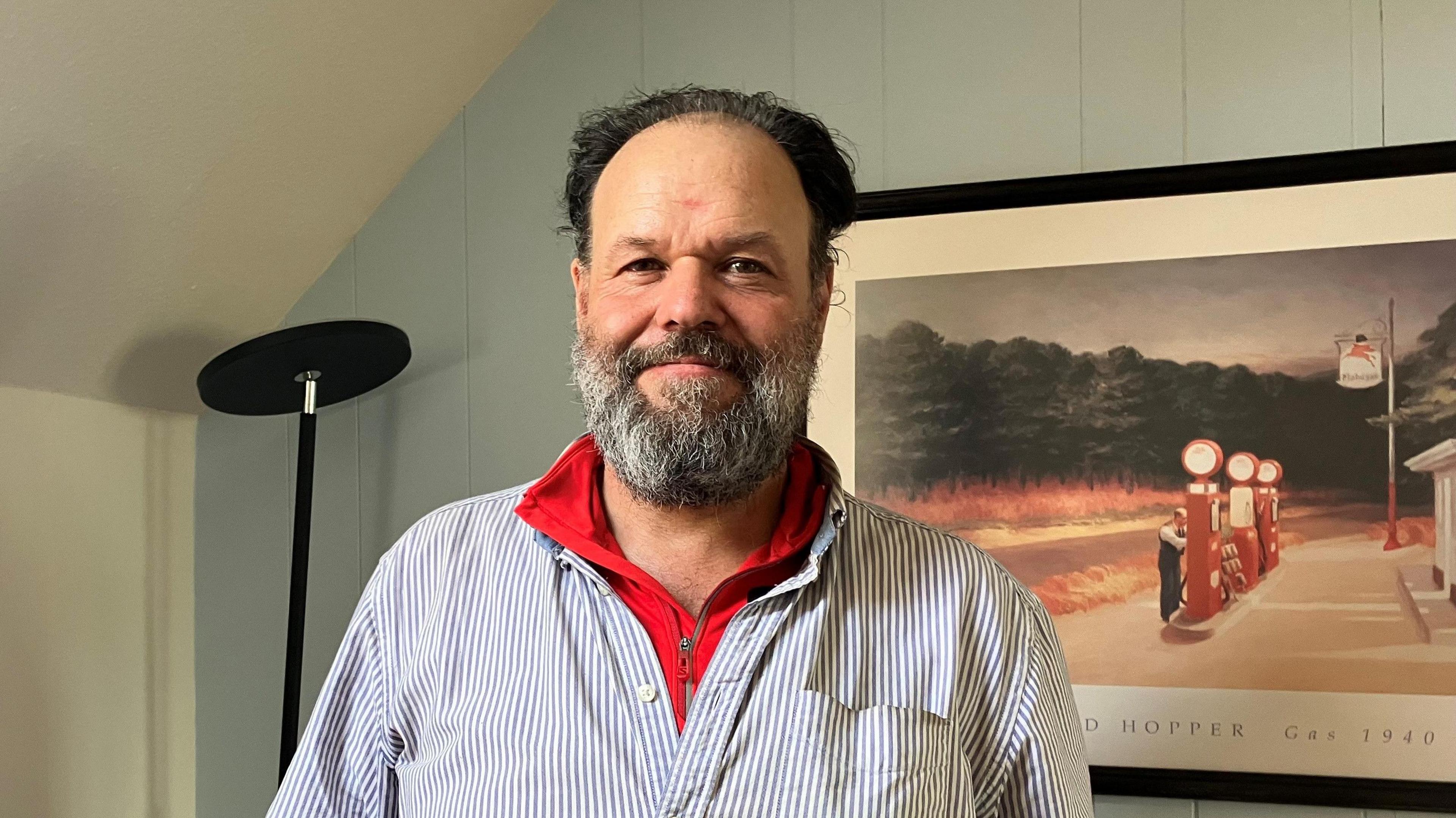 Sean Bright, wearing a red long-sleeved top and a blue and white striped shirt, stands in front of a painting in a room painted in a pale blue