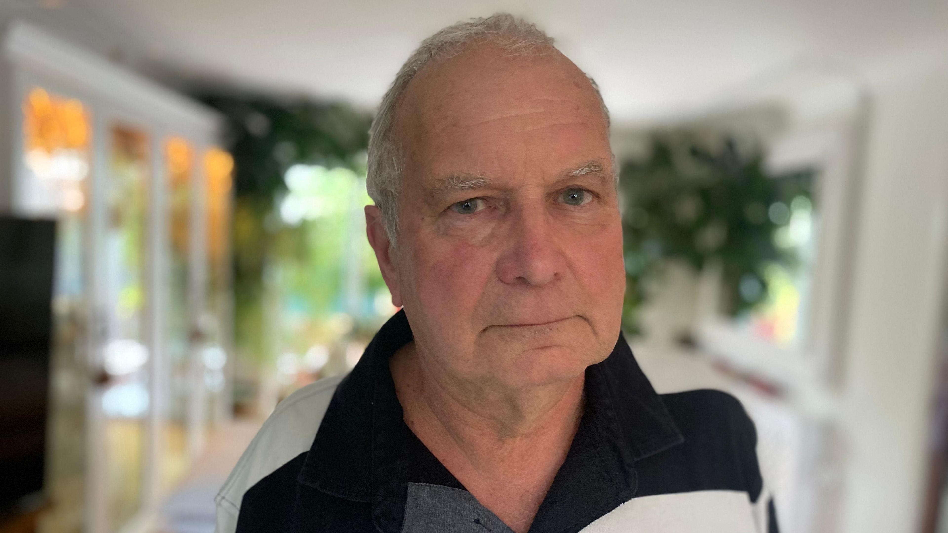 Clive Vaughan looks into camera. He is in his living room with plants behind him. He has short white hair and is wearing a black and white striped polo shirt.