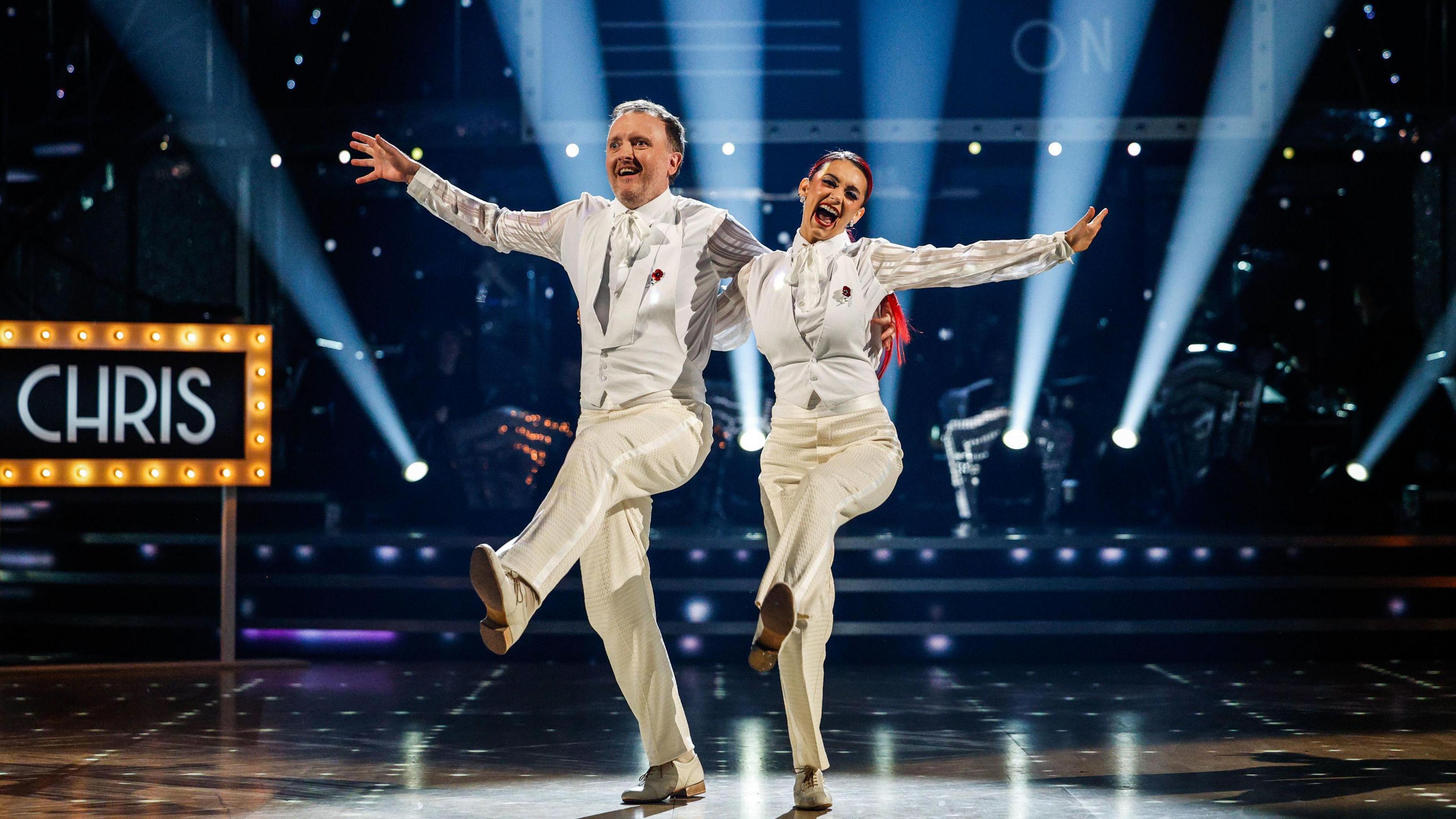 Chris and Dianne are smiling and wearing matching white suits on the dance floor, with an arm round each other. They are both kicking up their left legs in unison. The set is dark, with a follow-spot on the couple, and seven uplighters projecting white light upwards behind them. Chris's name is on a sign surrounded by light bulbs behind them.