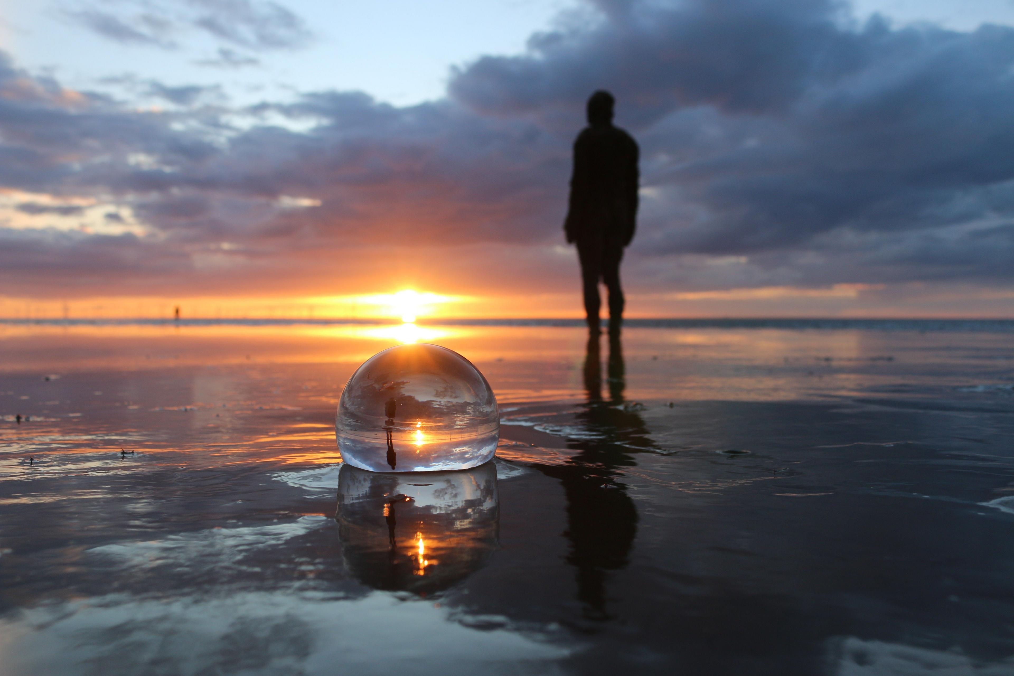 The figure of a male statue by artist Antony Gormley is reflected in a glass lens ball which has been placed in the sand on a beach at sunset.