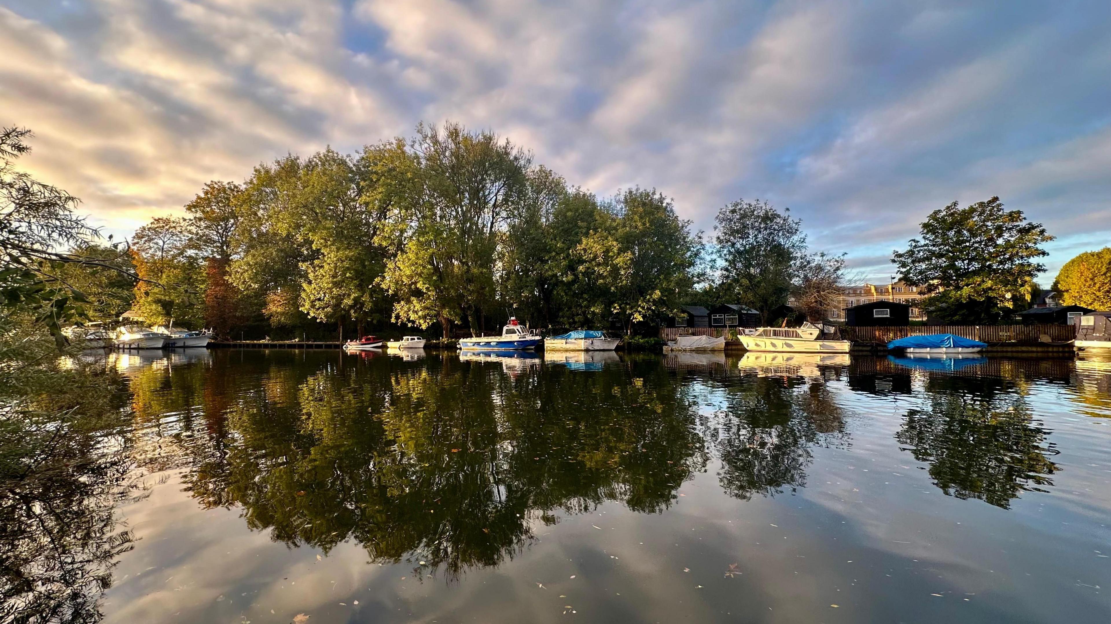 Streaks of sunlit cloud appear to spiral outwards from a cluster of trees beside a river with moored boats. The trees and cloud pattern are mirrored in the still water.
