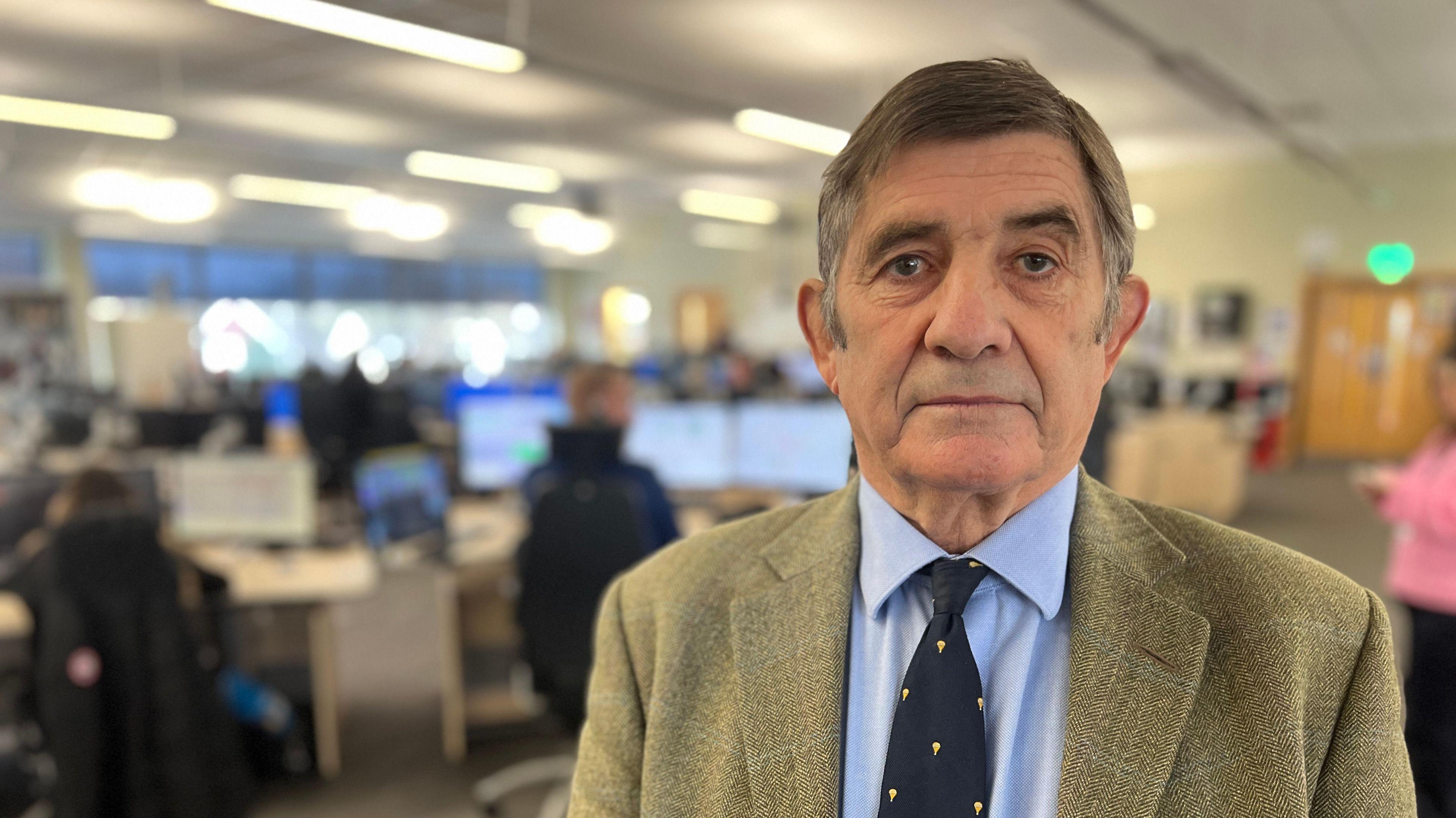 Philip Wilkinson looks at the camera. He is in a busy police office area. He wears a blue shirt and tie with a tweed jacket.
