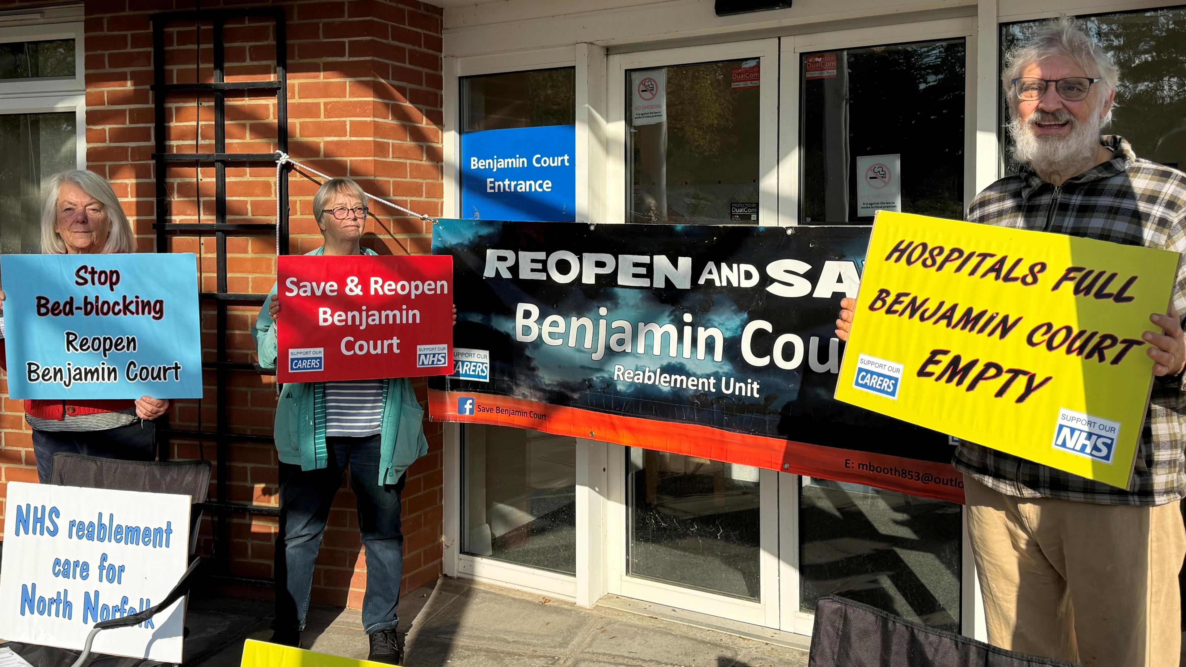 A group of three campaigners outside a former NHS building, known as Benjamin Court in Cromer. They are all standing and looking towards the camera and are holding signs. Some of the signs read, 'NHS reablement care for North Norfolk', 'Stop bed-blocking Reopen Benjamin Court', 'Save & Reopen Benjamin Court' and 'HOSPITALS FULL BENJAMIN COURT EMPTY'.