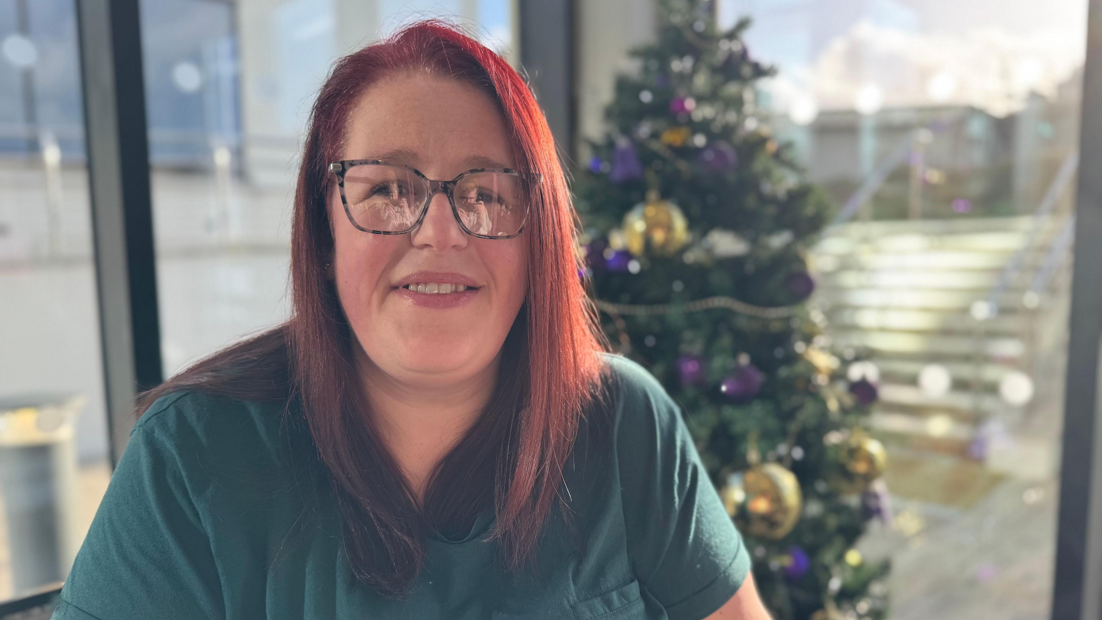 A women with long red hair smiling at the camera. She has patterned glasses on. She is wearing a dark green t-shirt. There is a Christmas tree behind her left hand side.