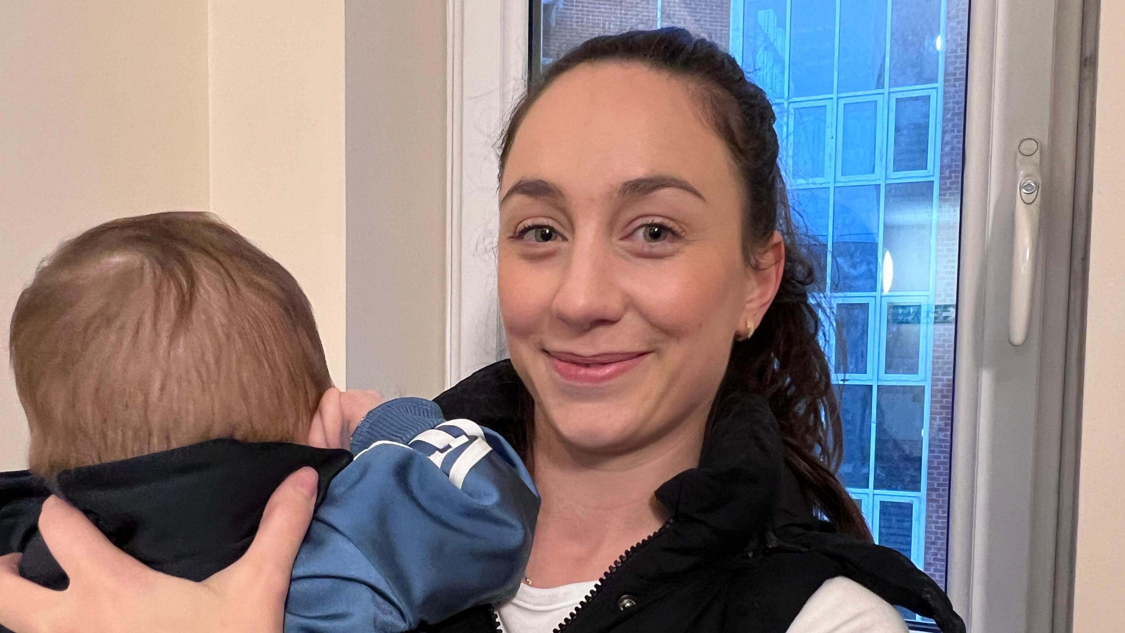 Woman holding baby in front of a window. Behind you can see a block of flats. 