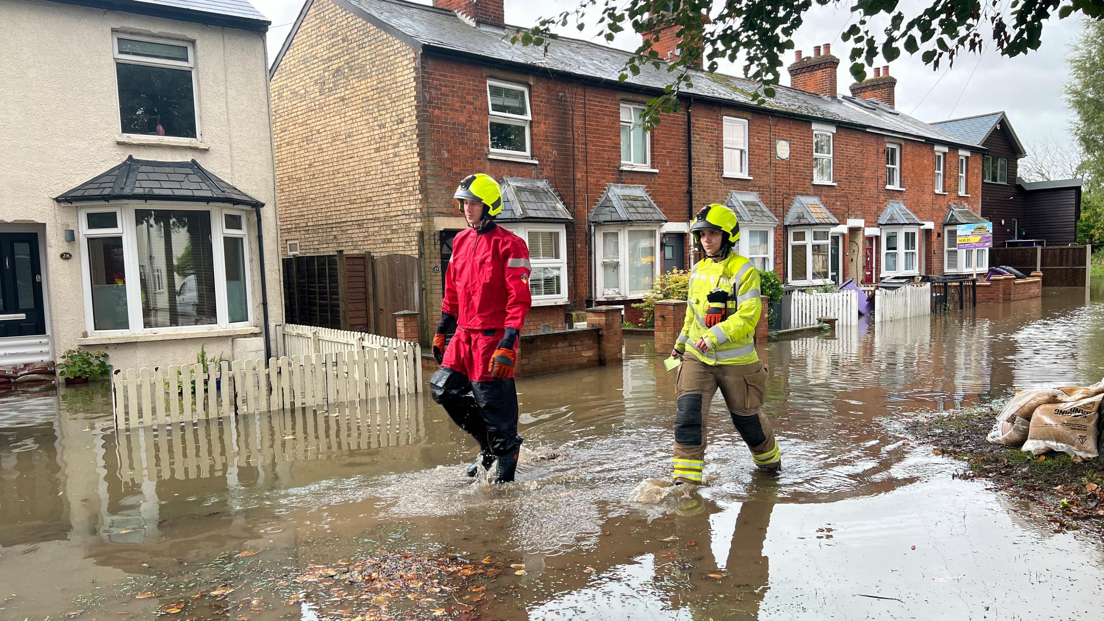 Two firefighters wearing helmets and flood gear wading through a flood outside housing 