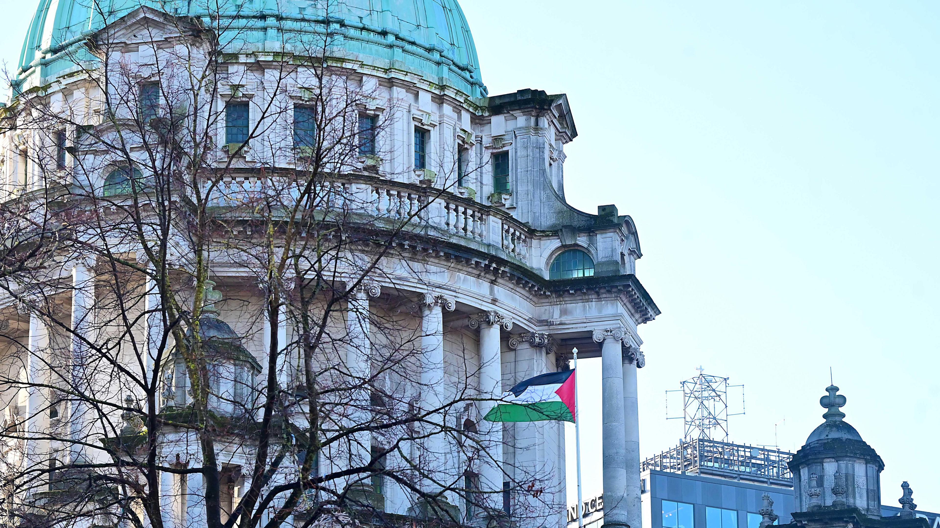 The Palestine flag is flapping in the wind outside a baroque revival style building. Several pillars decorate the building. The roof is a green dome. The sky is light blue. A tree is bare of leaves.