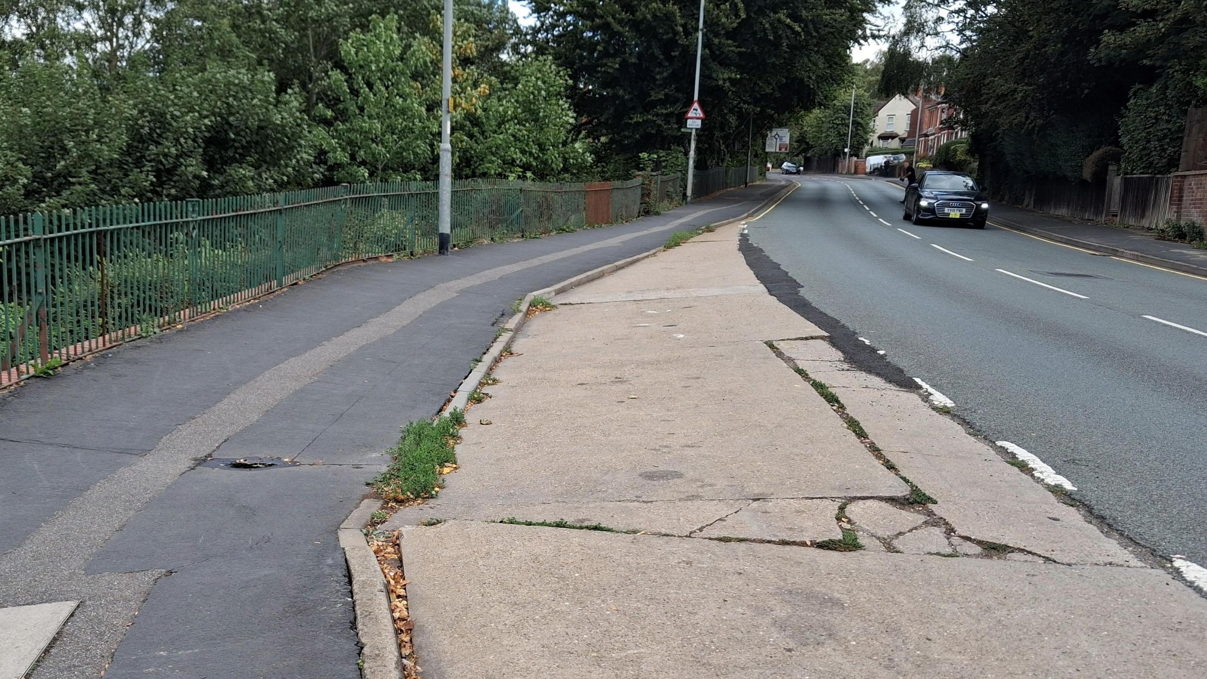 View of a road on an incline. There are patches of tarmac where repairs have been carried out and a green railing along the side of the path. There are several vehicles travelling down the hill, including a black Audi.