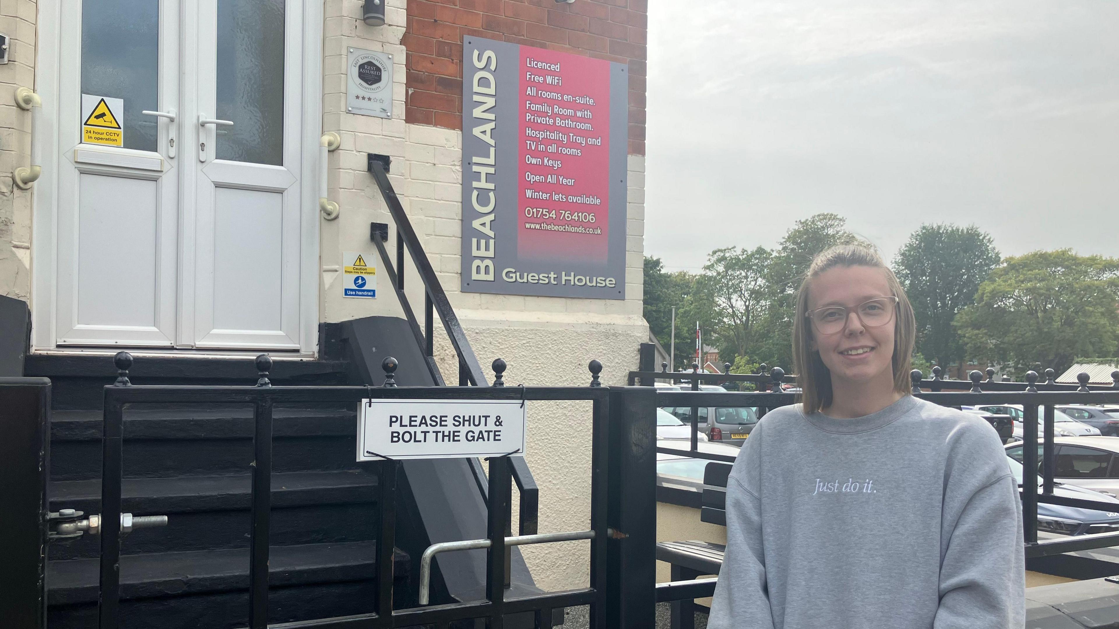 A woman with shoulder-length dark blonde hair and a grey hoodie that says 'just do it' is stood next to a black gate. Next to the gate are steps leading up to two white doors. A sign to the right of the door says 'Beachlands Guest House' with a car park to the right of it.