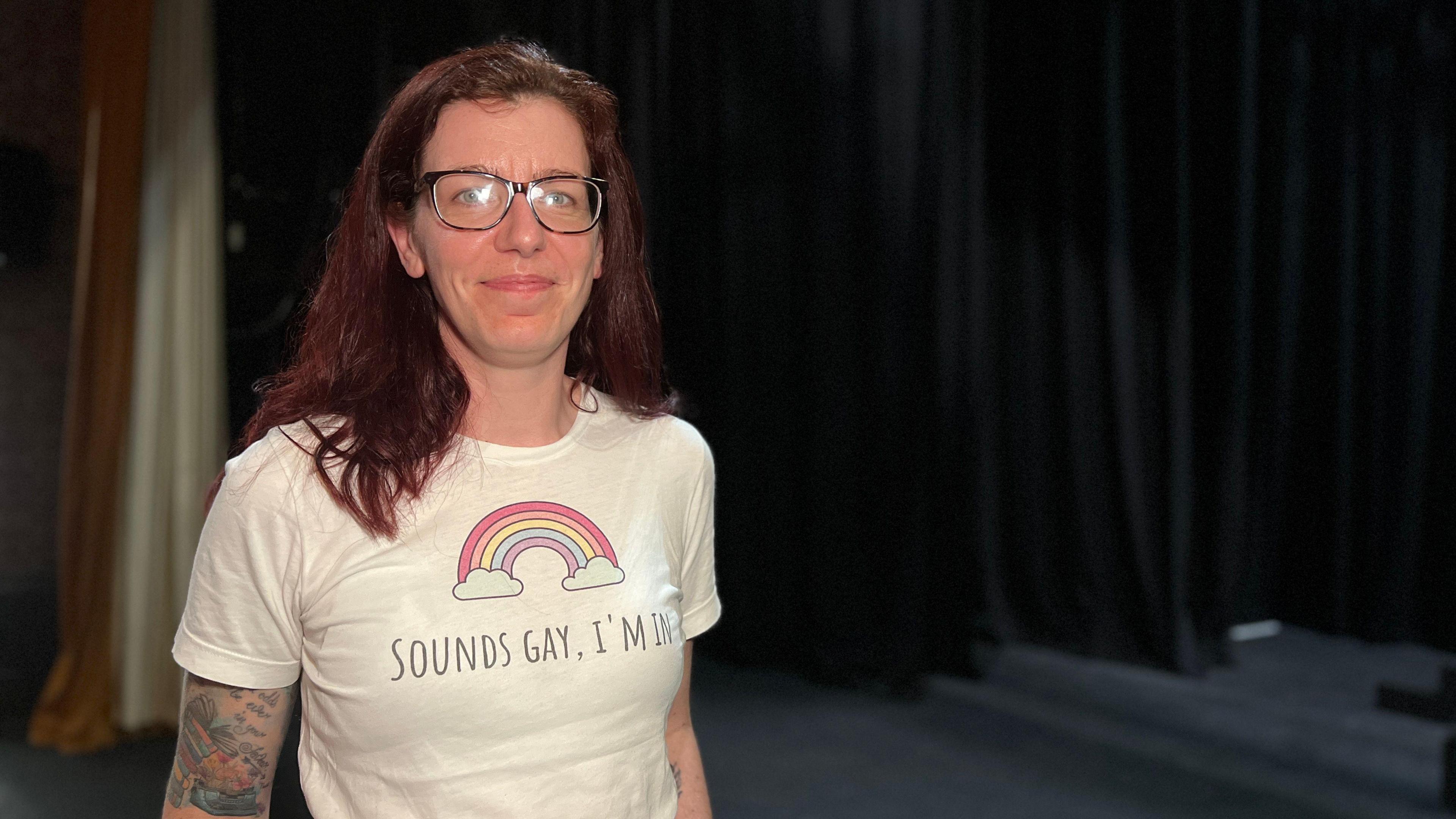 Kaye smiles at the camera with a white t-shirt on with a rainbow on it and text saying 'Sounds Gay, I'm In'. She has dark red hair which comes down past her shoulders and is wearing glasses. Behind is are the black stage curtains.