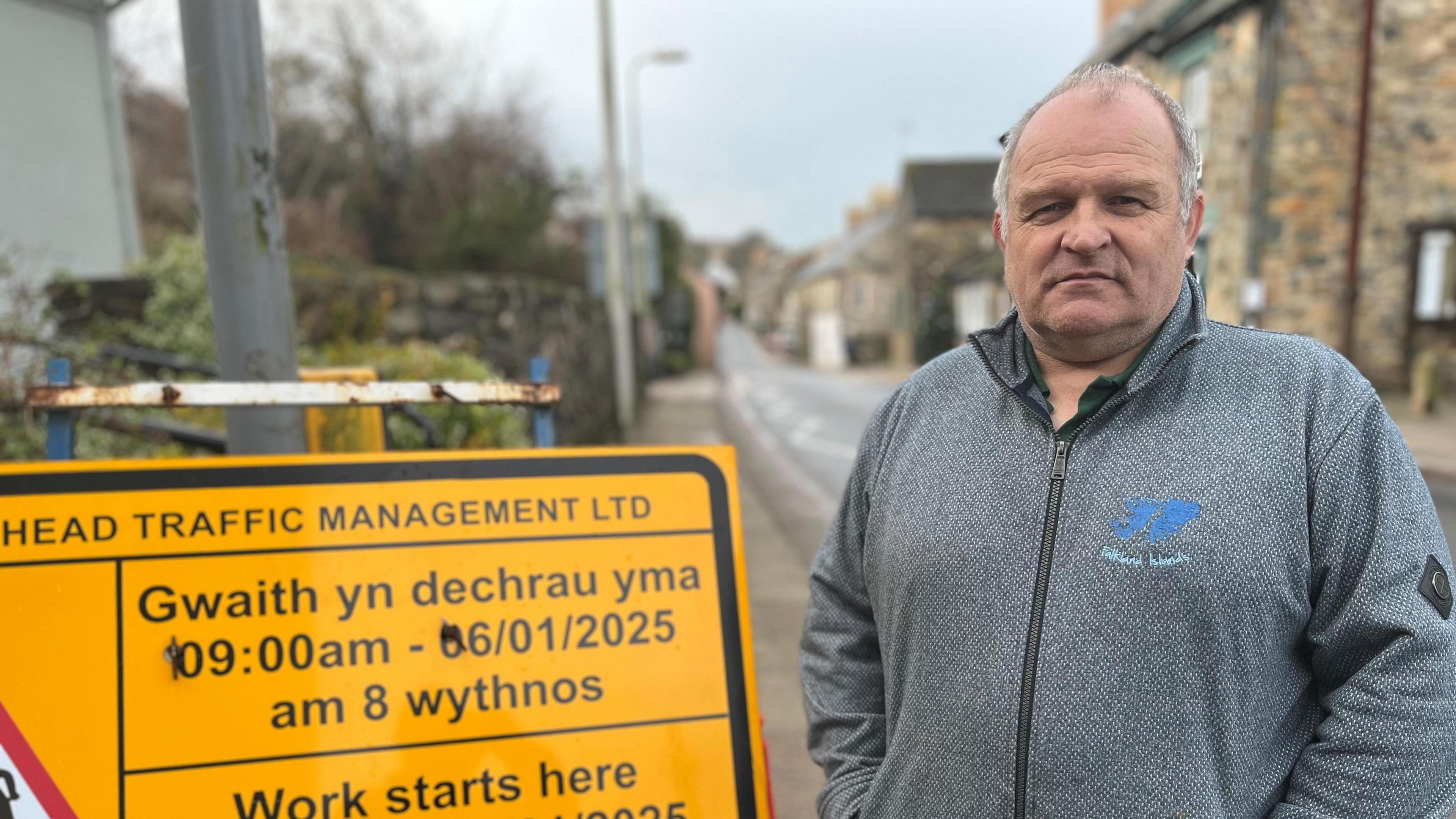 Huw Murphy is wearing a grey zip-up fleece and has short grey hair. He is stood next to a yellow road sign which reads "gwaith yn dechrau yma 09:00 - 06/01/2025 am 8 wythnos", which is Welsh for "work starts here at 09:00 - 06/01/2025 for eight weeks"