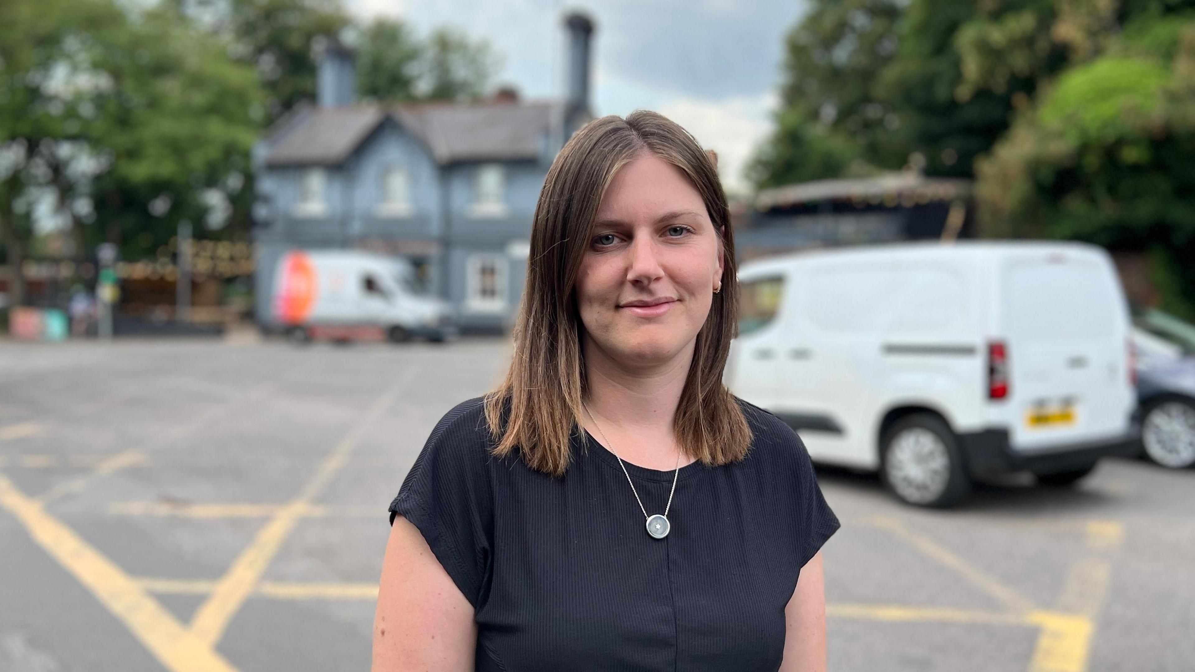 Katy Goldsmith stands at the road junction. The Last Crumb pub is visible in the background and a yellow-box junction can been seen on the road surface behind her. A white van is in the process of making a right turn.