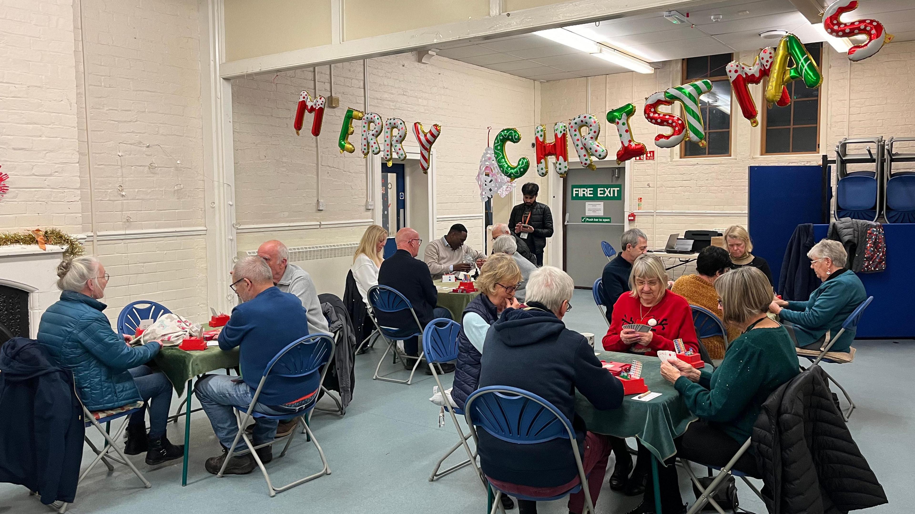 A room with four bridge tables set up with people sat around them. It's a village hall-type room, with a big banner reading "Merry Christmas" spelled out by brightly-coloured balloons. 