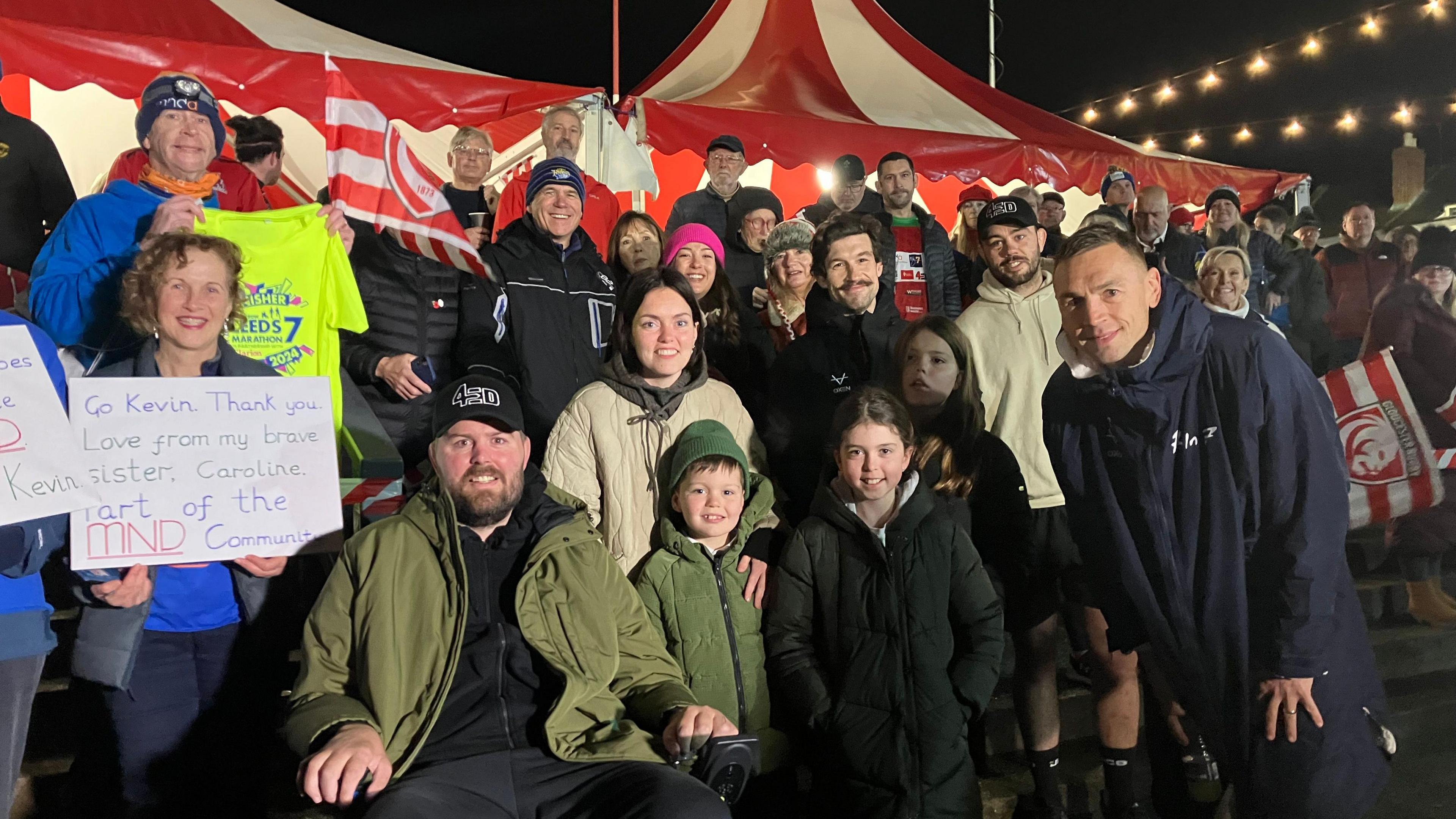 Kevin Sinfield, right, wearing a long navy coat, smiling with supporters before leaving Kingsholm. There is a large red and white circus-style tent in the background and a large group of people smiling at the camera. 