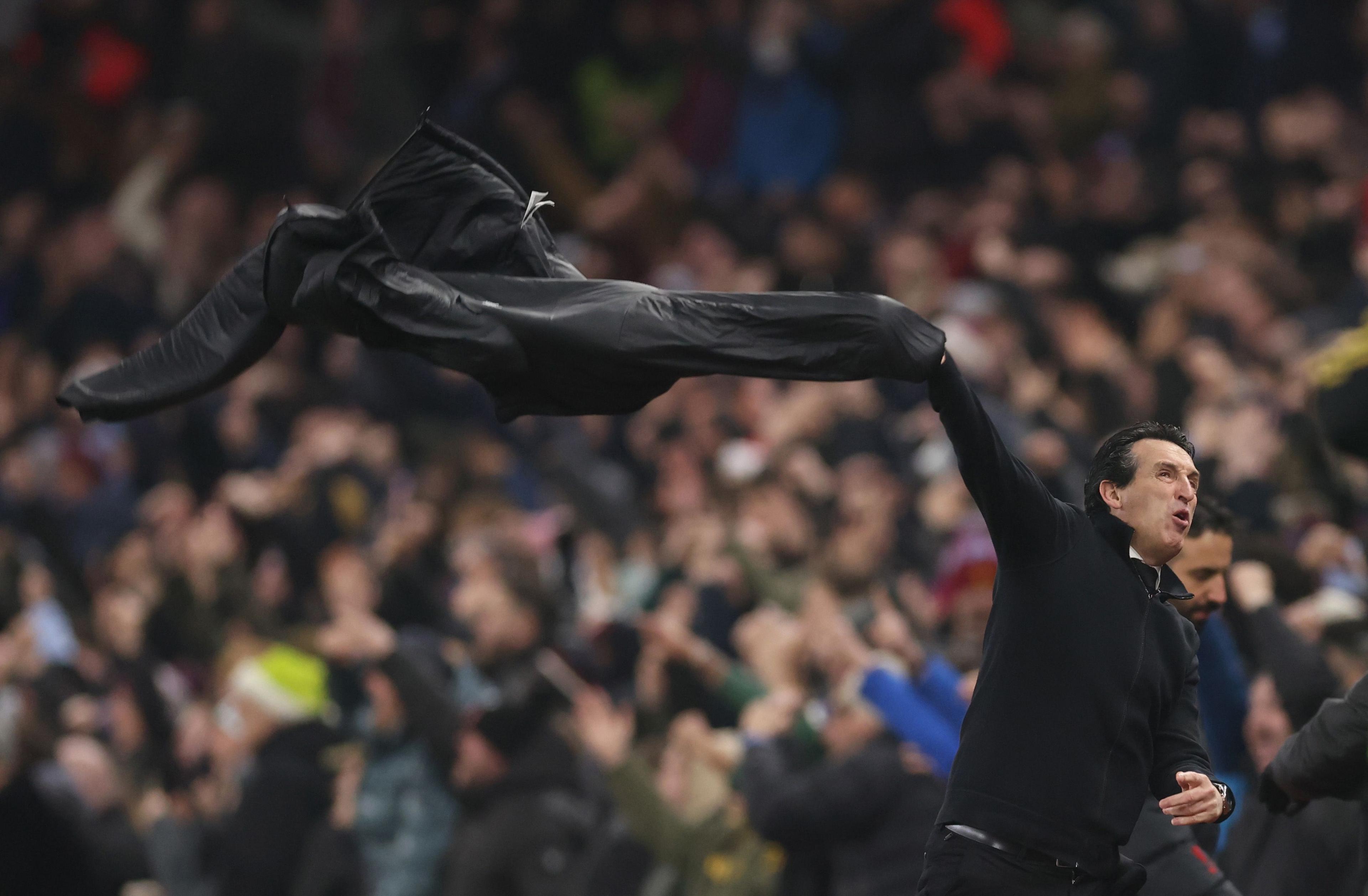 A person on the touchline swings a black jacket dramatically through the air in a moment of intense celebration, with a packed stadium crowd cheering in the background.