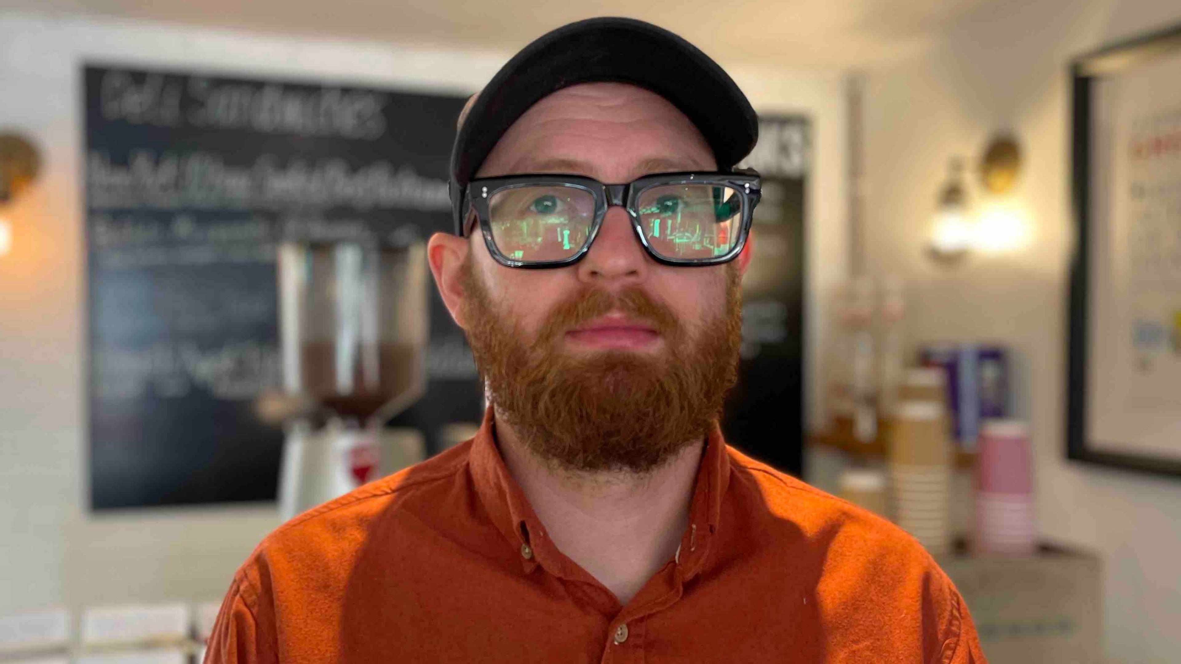 Tom Ertman who runs the Cheddar Gorge deli, pictured in his shop. He has a ginger beard, heavy black-rimmed glasses, a baseball bap and is wearing an orange shirt.