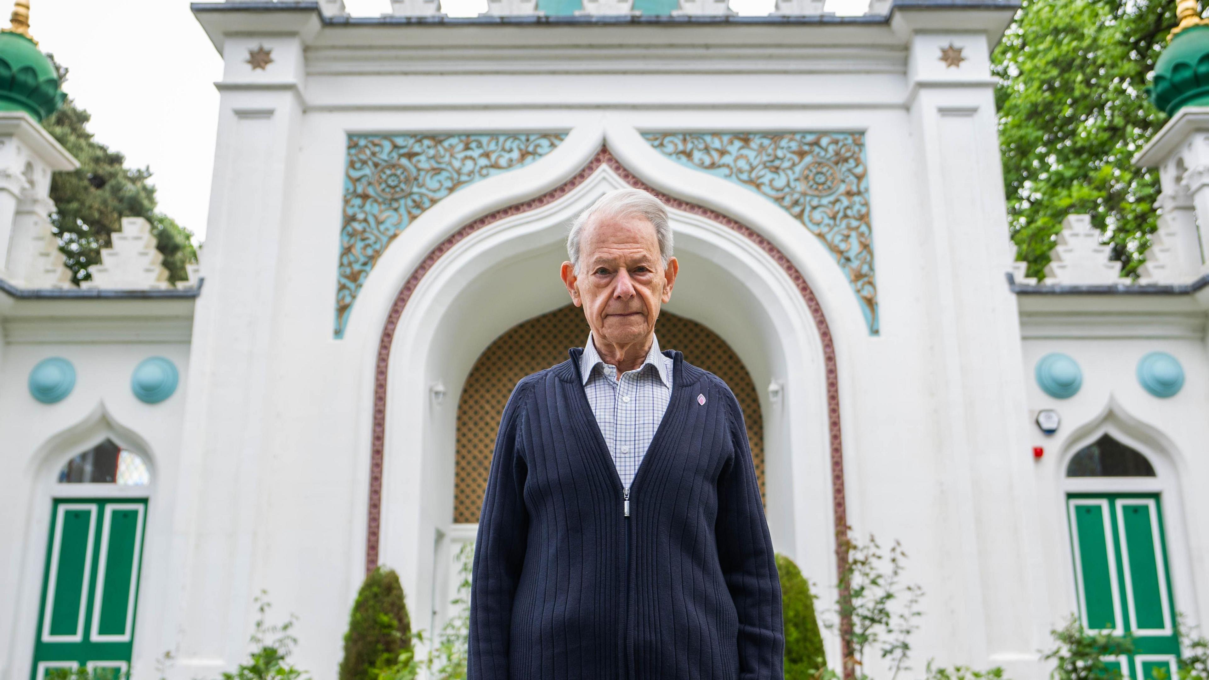 An elderly man wearing a grey shirt and dark blue cardigan. He is facing the camera and stood outside a white mosque