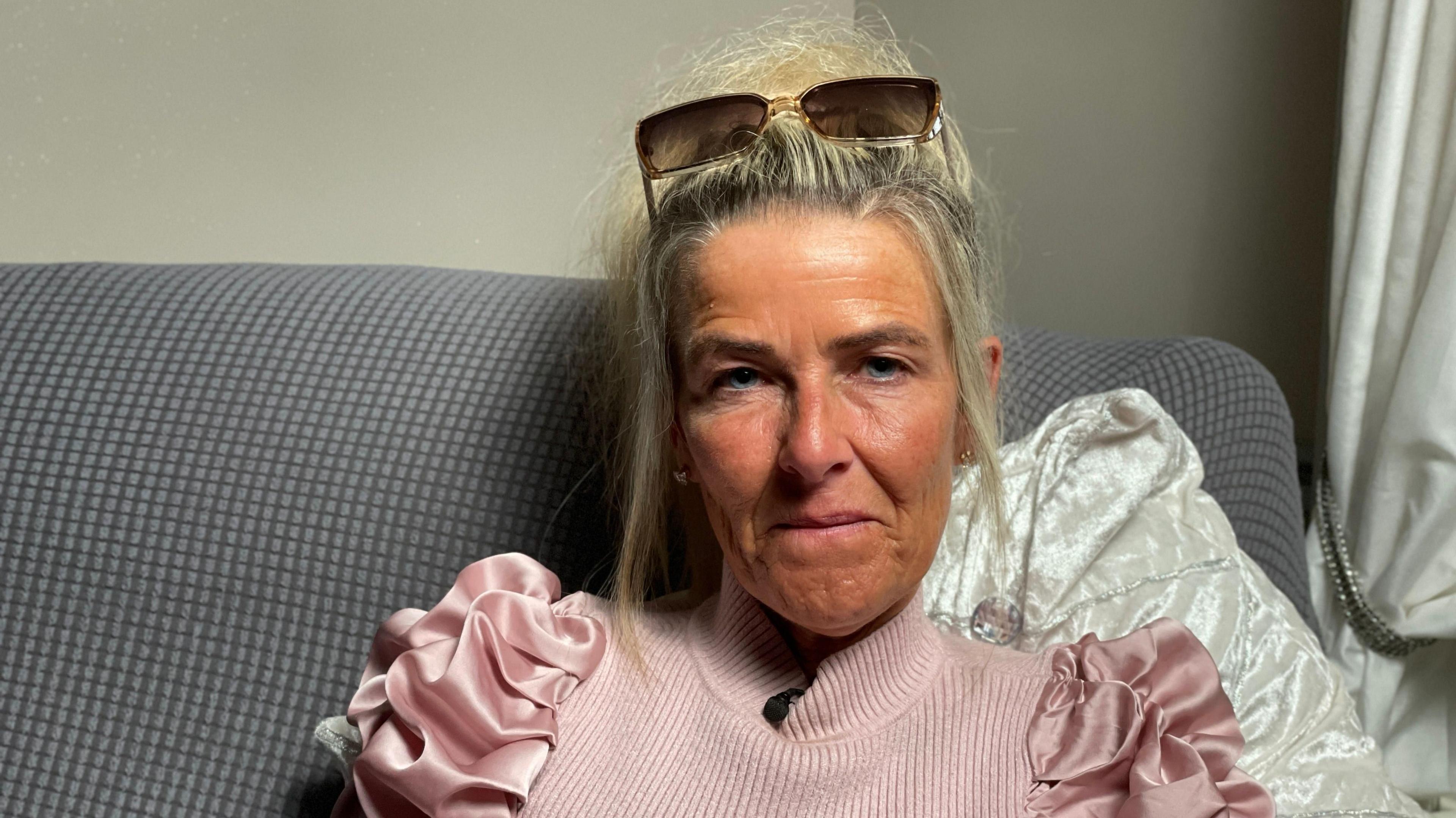 A woman wears a pink jumper and sunglasses on her head, sitting on a grey sofa looking at the camera.