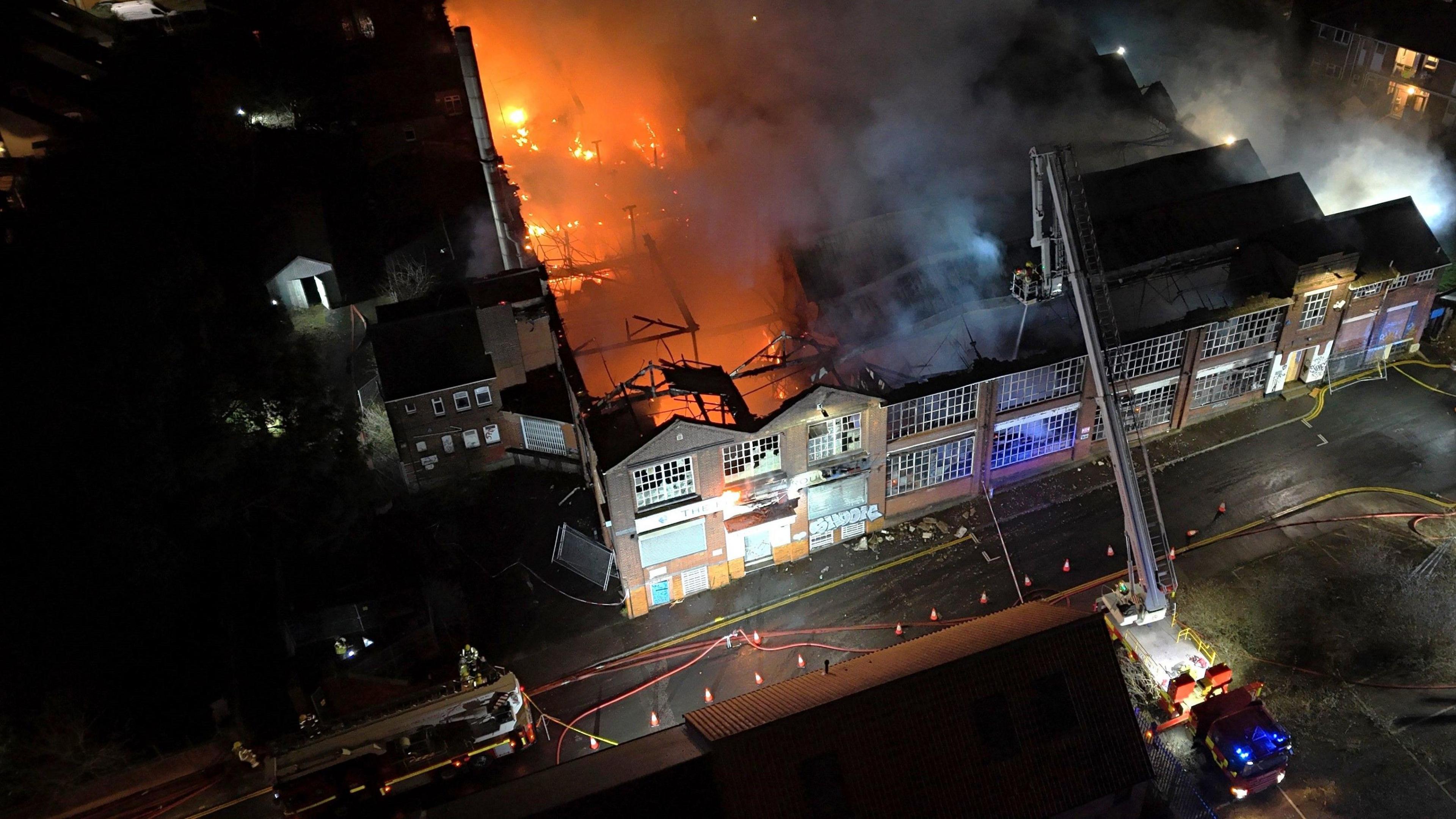 A fire service aerial platform ladder extinguishing the blaze. Half of the building is fully alight with flames and smoke filling the air