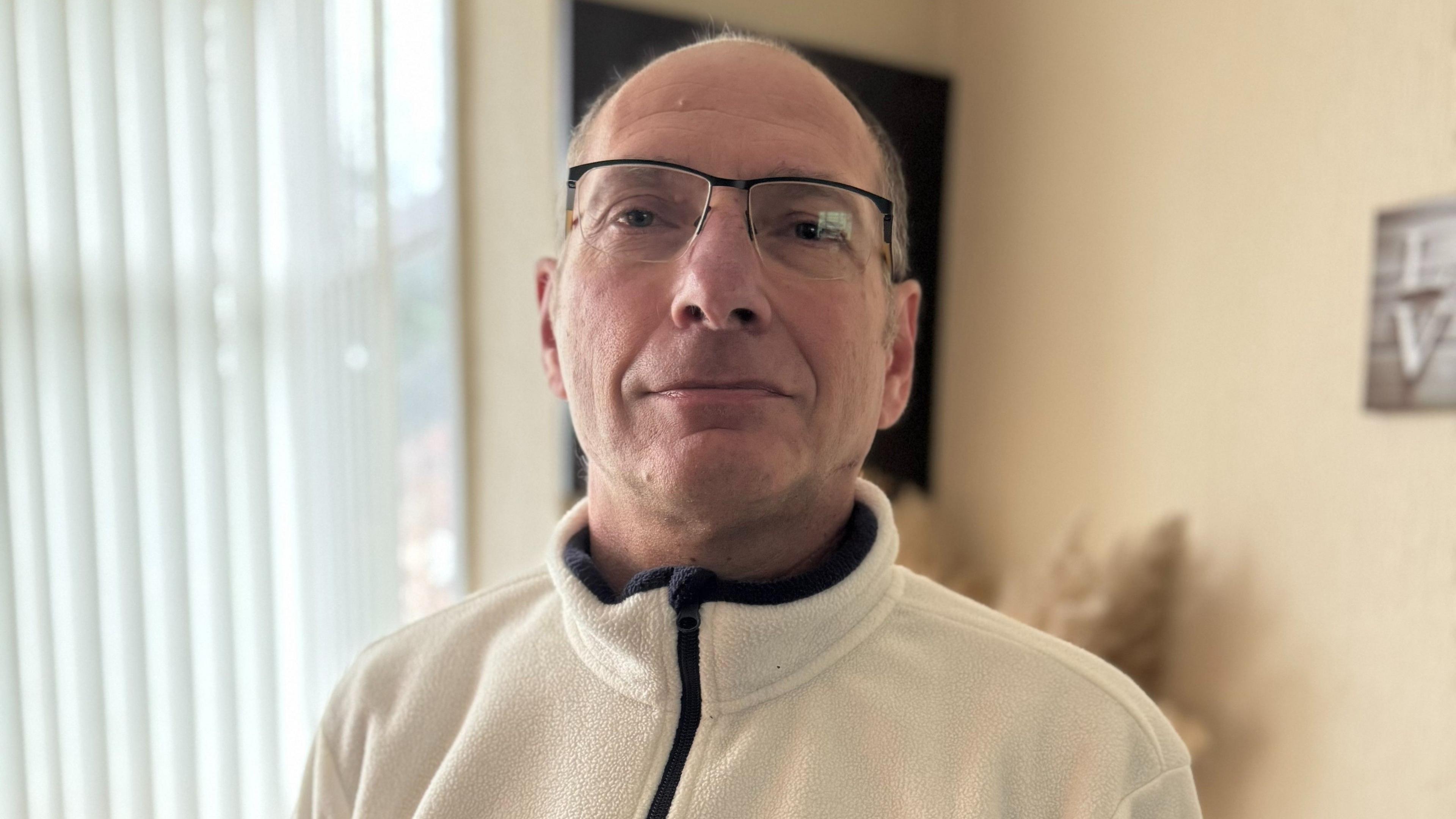 A man stands looking into the camera. He is balding and is wearing glasses and a white fleece top with a zip. Behind him is a beige wall and window with blinds.