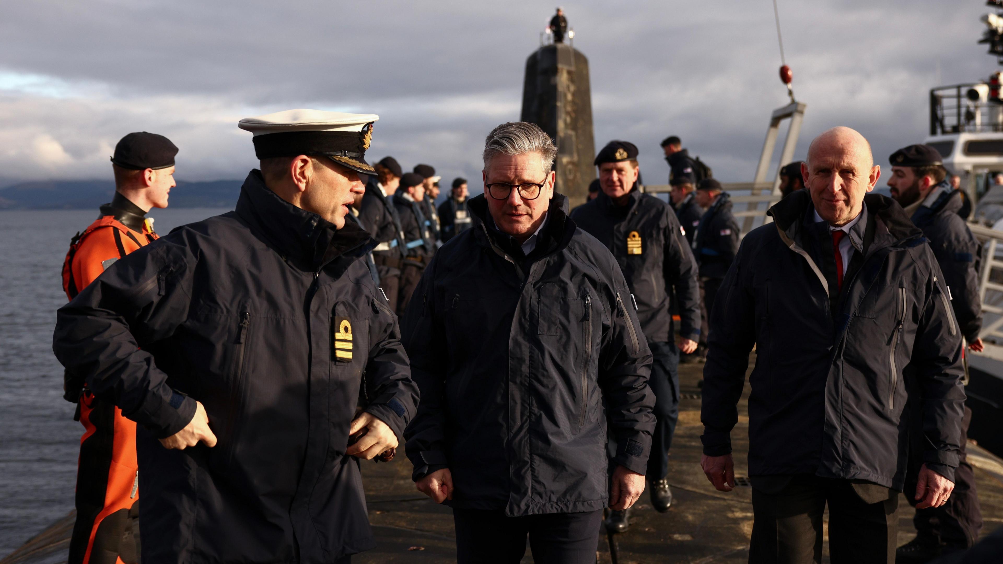 Prime Minister Sir Keir Starmer and Defence Secretary John Healey visited a Vanguard-class submarine in March. They are on board the deck in thick parkas. A naval officer in full outdoor uniform is speaking to them.