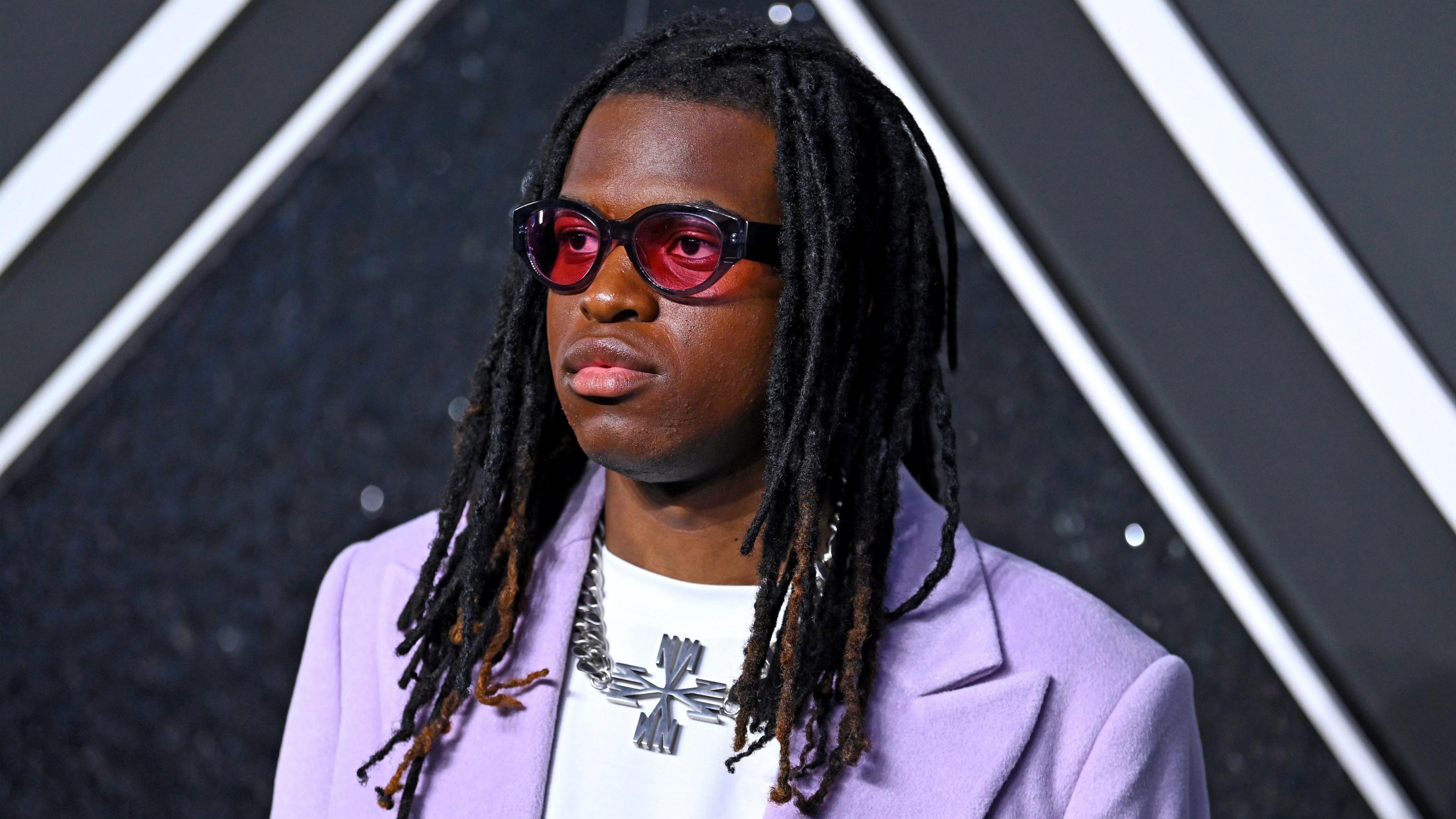 Jordan Adetunji attending an event. He's getting his photo taken by press. He's wearing sunglasses and a purple suit with a white top and a silver neckace. 