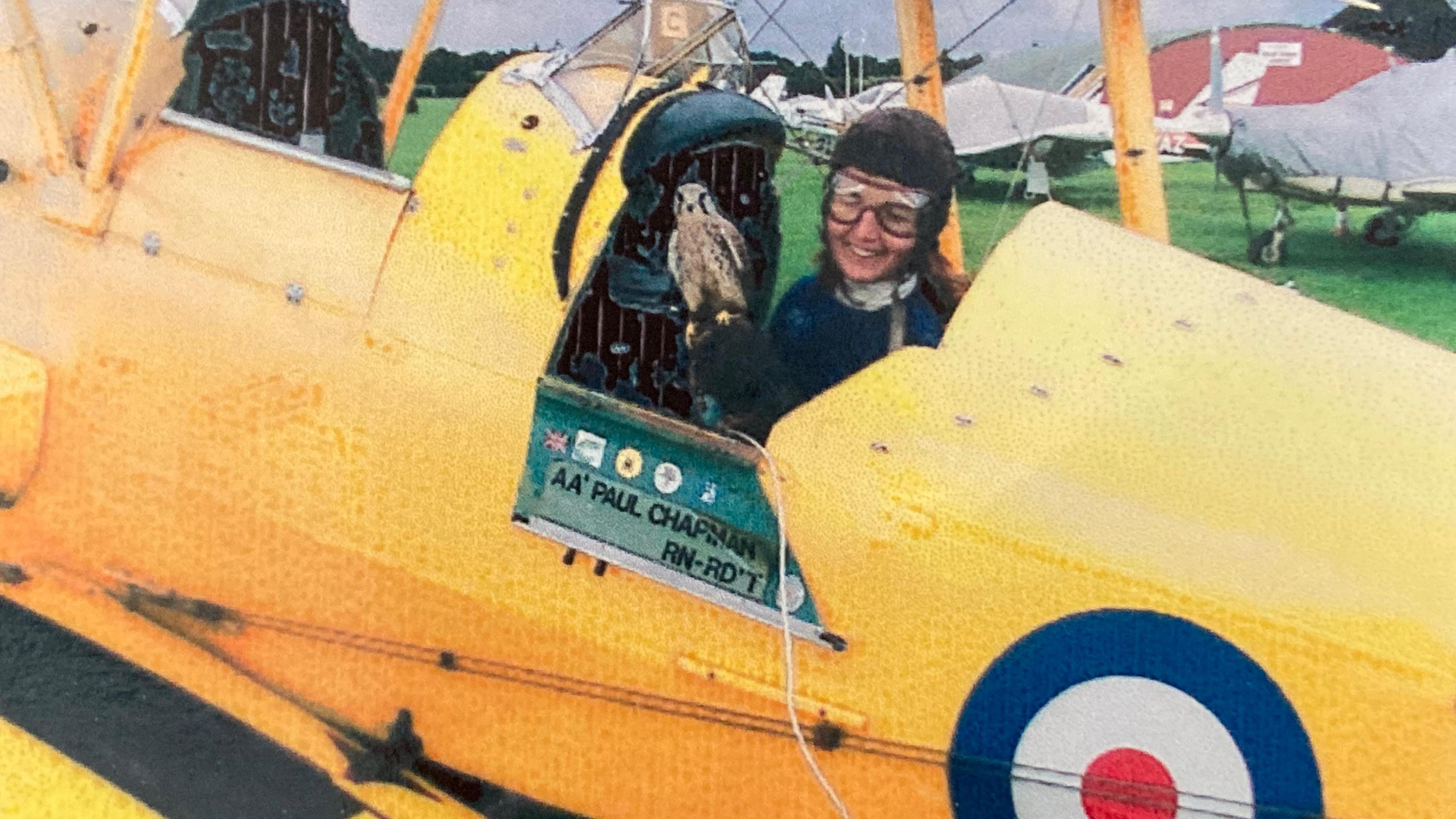 Tizi Hodson smiles from the pilot's seat of a yellow biplane with an RAF roundel on the side. She is wearing goggles and a pilot's helmet. The plane is parked at an airfield with other planes and a hanger in the background.