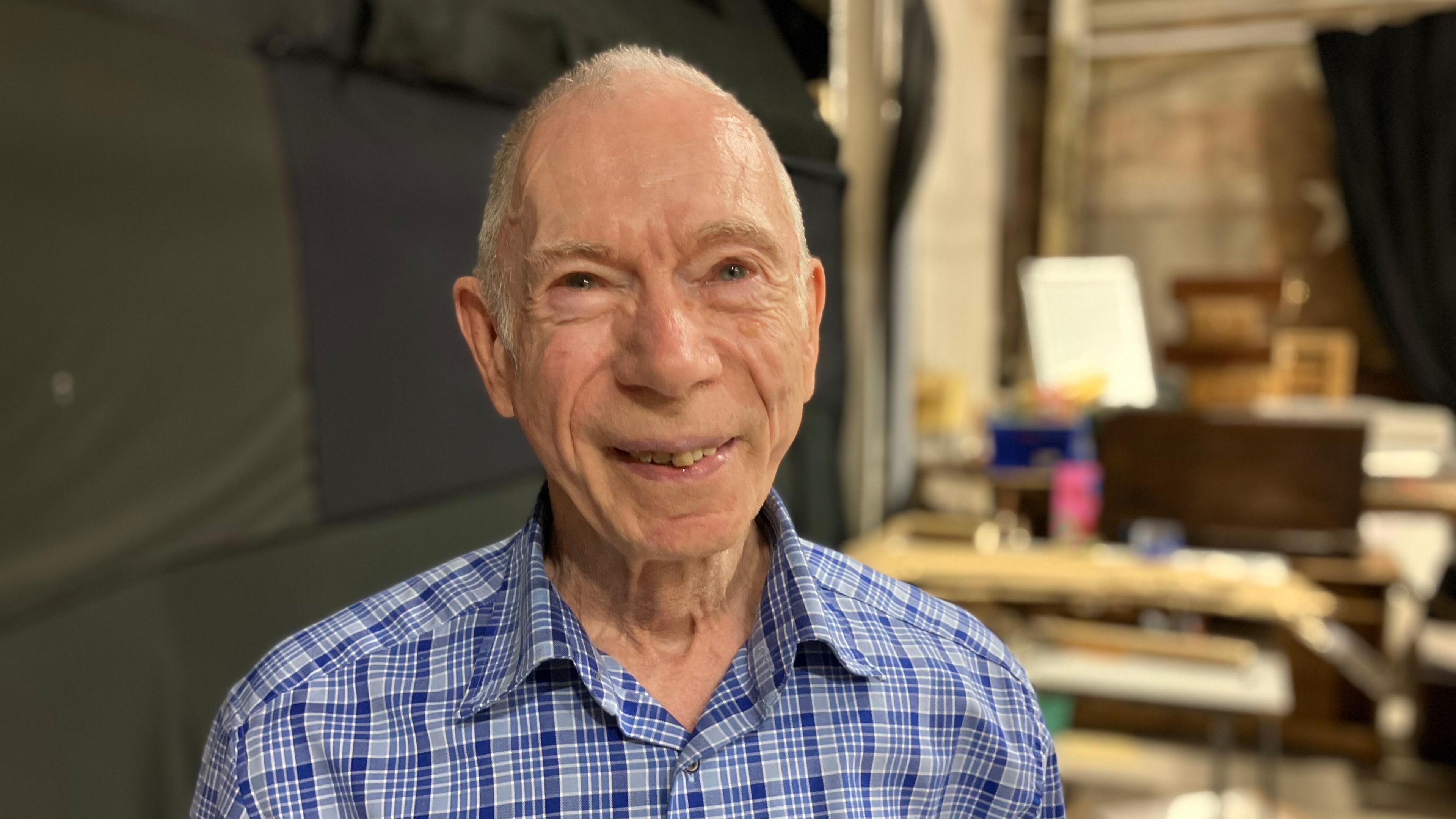 An man with cropped white hair looking towards the camera in a workshop or backstage area. He has a blue chequered shirt on.