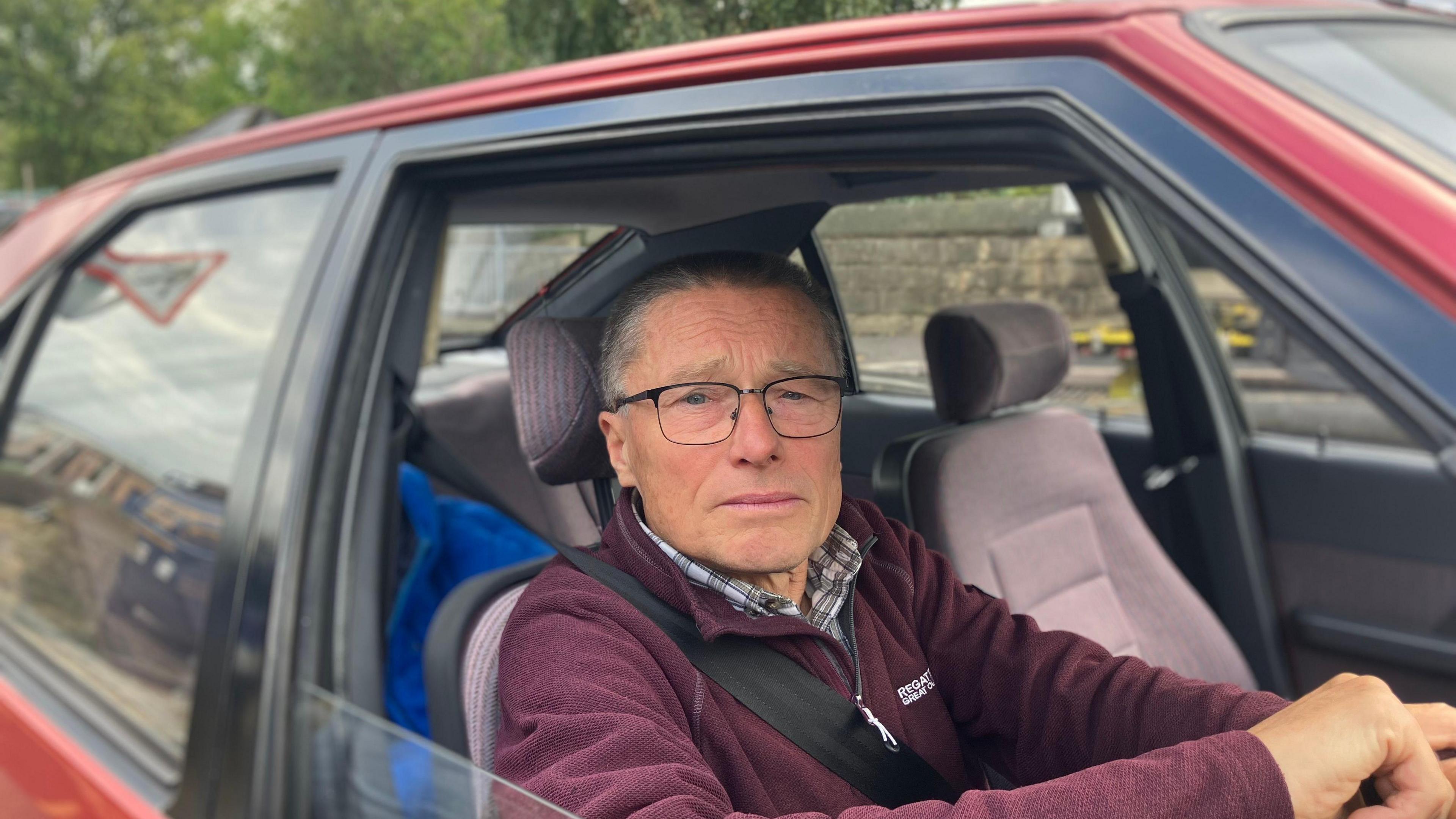 Andrew sits in a car dressed in a burgundy fleece waiting for his son