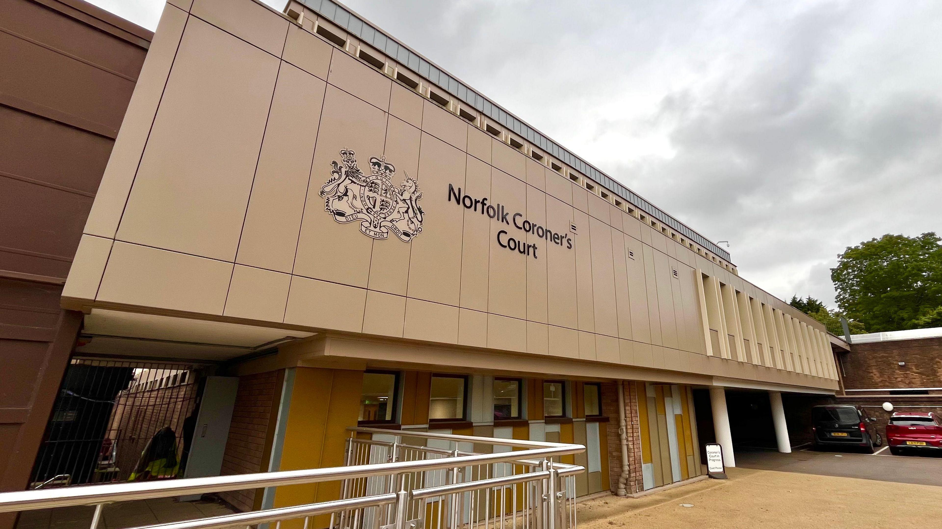 The front of Norfolk Coroner's Court. The building is brown, and has a big logo and title on the front. At the bottom, you can see railings and a walk way. On the right, two cars are parked. 