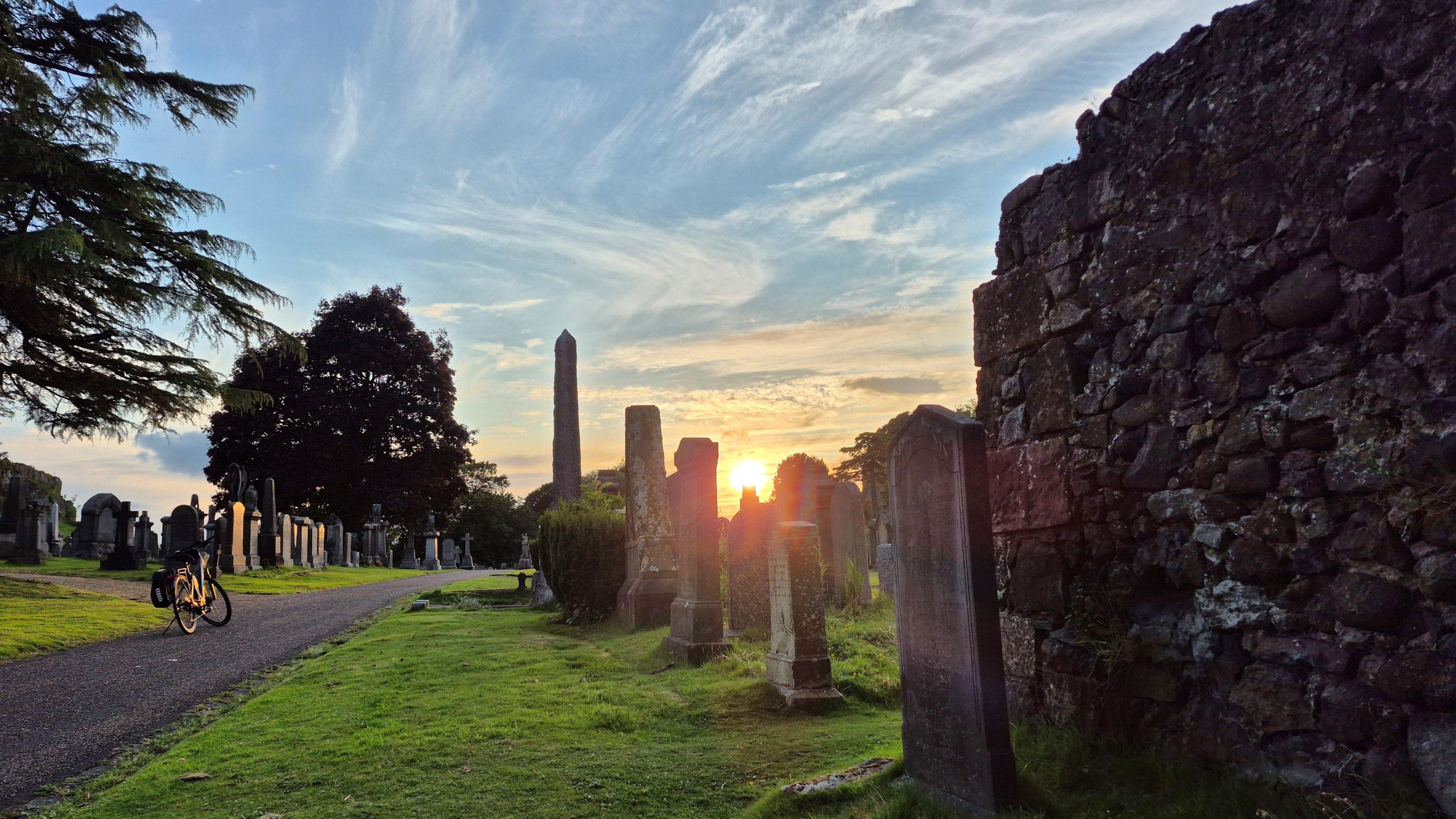 A cemetery during golden hour. Several dark-coloured gravestones run up the middle of the image. The sun can be seen going down between the middle two. The sky around it is orange and the rest of the sky is light blue and streaked with white cloud. Two areas of green grass are separated by a grey path running through it, where a bike is parked.