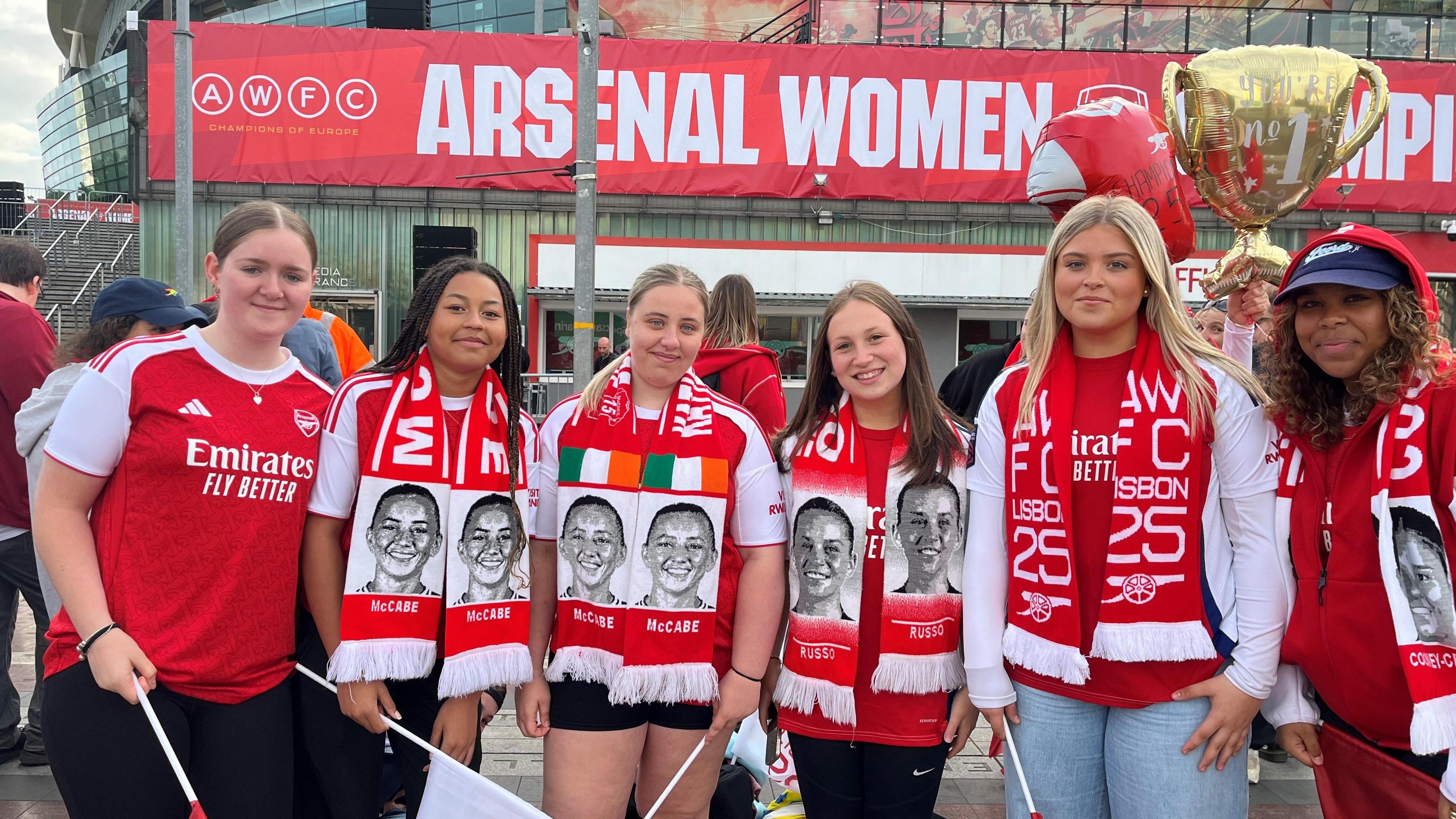 Teenager fans Talula, Amelia, Maddie, Scarlett, Charlotte and Taeja stand outside the Emirates Stadium looking at camera, dressed in Arsenal kits and scarves.