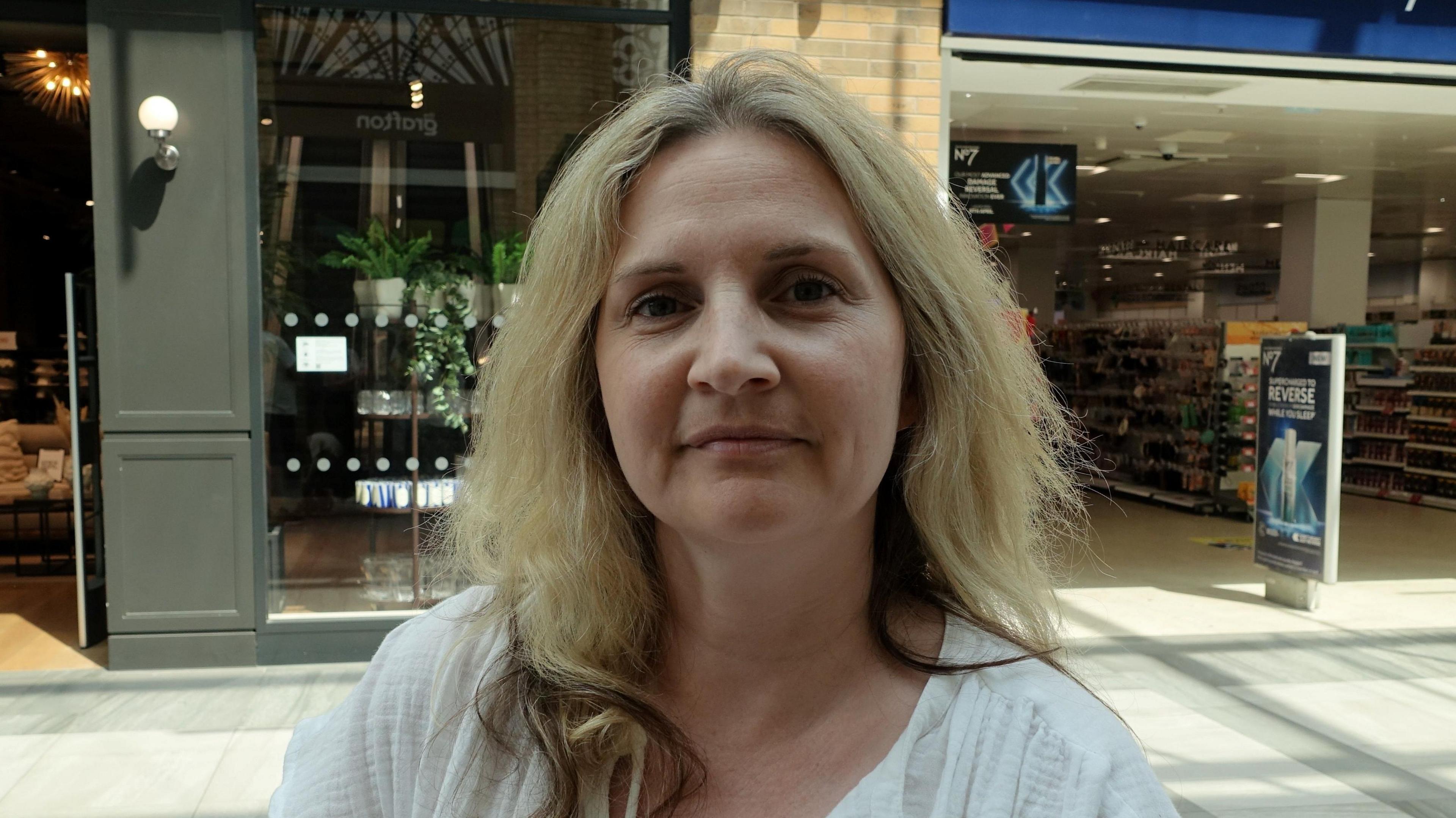 Head-and-shoulders photo of Sarah Lefever wearing a white top, with blonde hair, and shops in the background.