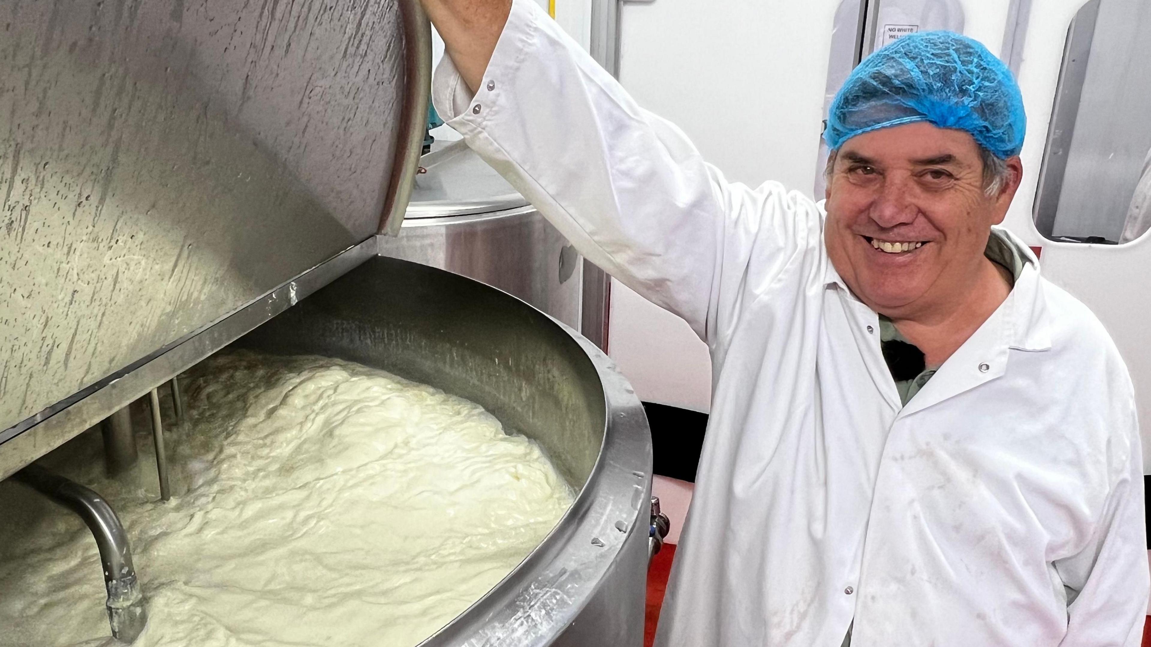 David Baker, wearing a white food production coat and blue hairnet, smiles as he lifts the lid on a steel vat, with frothy milk inside nearly to the top.