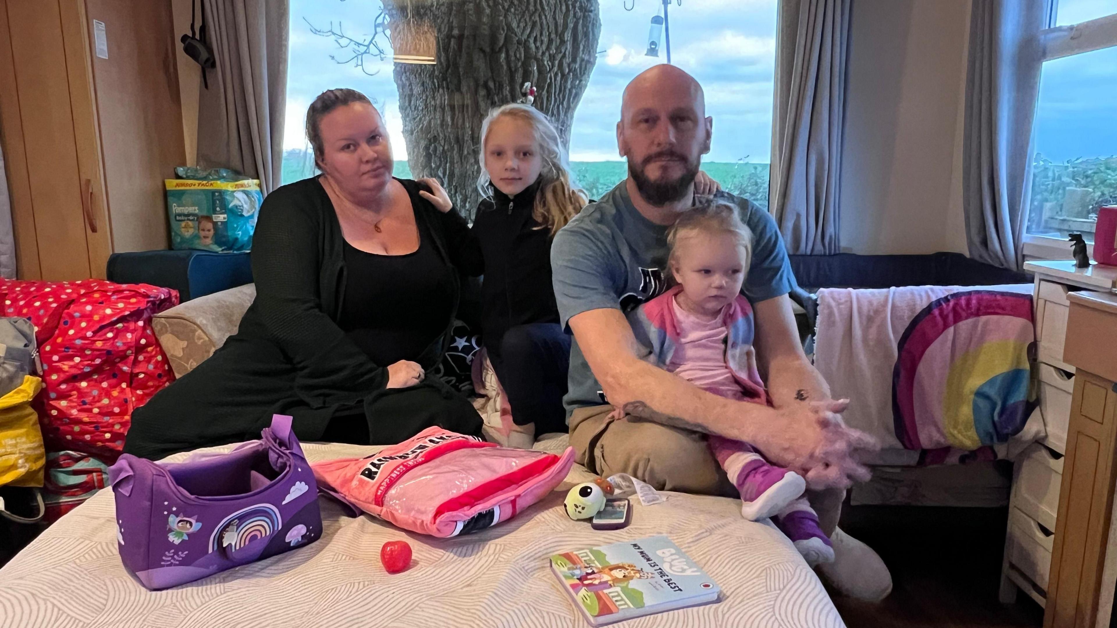 A family of four sat on a sofa bed in a caravan. There is a lot of stuff piled up in the corner to their left including a bag of nappies. A baby's cot is wedged to the right of the sofa. On the bed the mum, dad and two daughters sit, one of the girls is a toddler.  There is some toys and books on the bed with them.