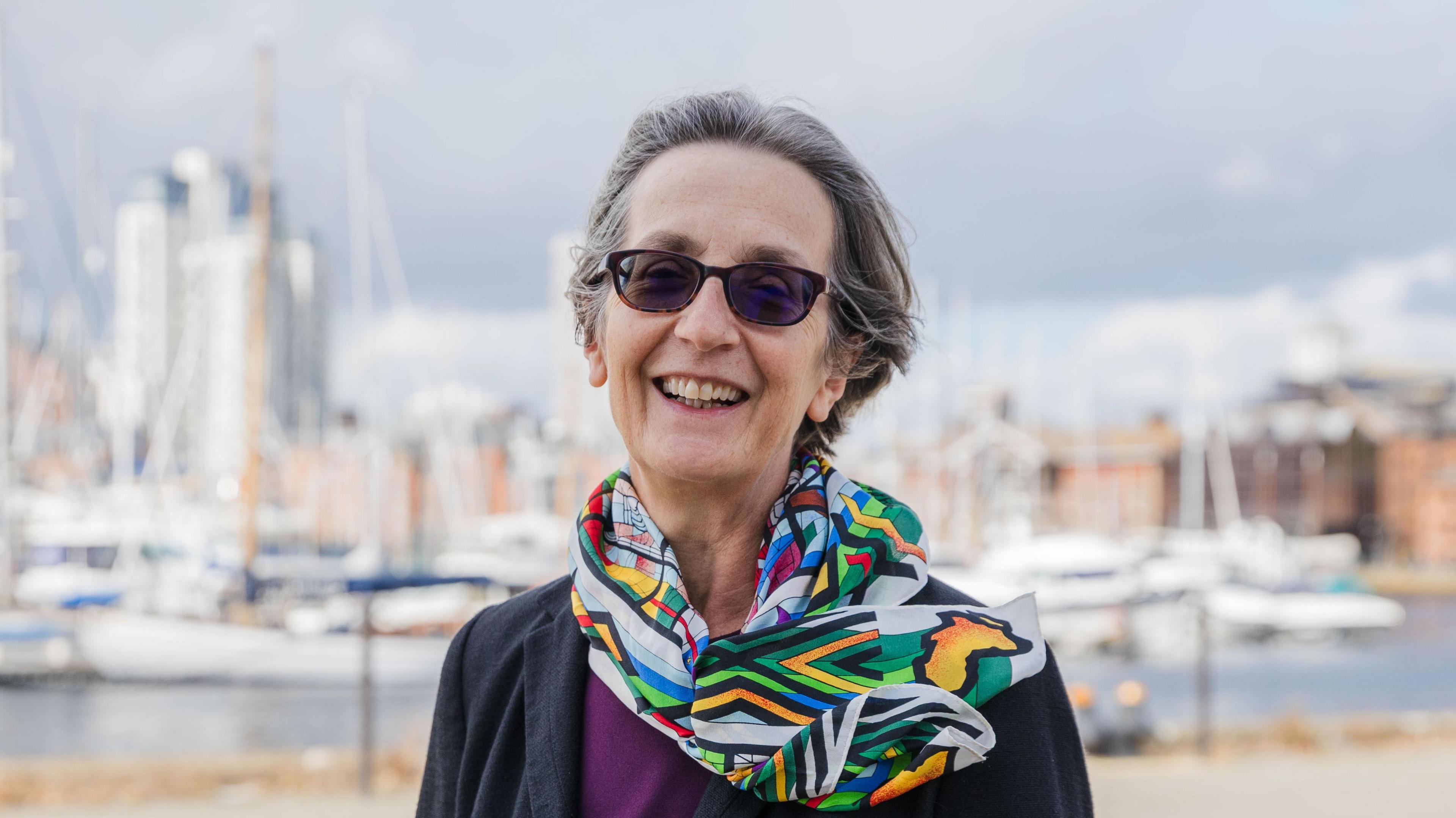 A head and shoulders image of Dr Helen Pankhurst CBE. She is wearing a colourful scarf and sunglasses. She is smiling and a standing in front of a blurred back of boats on a waterfront.