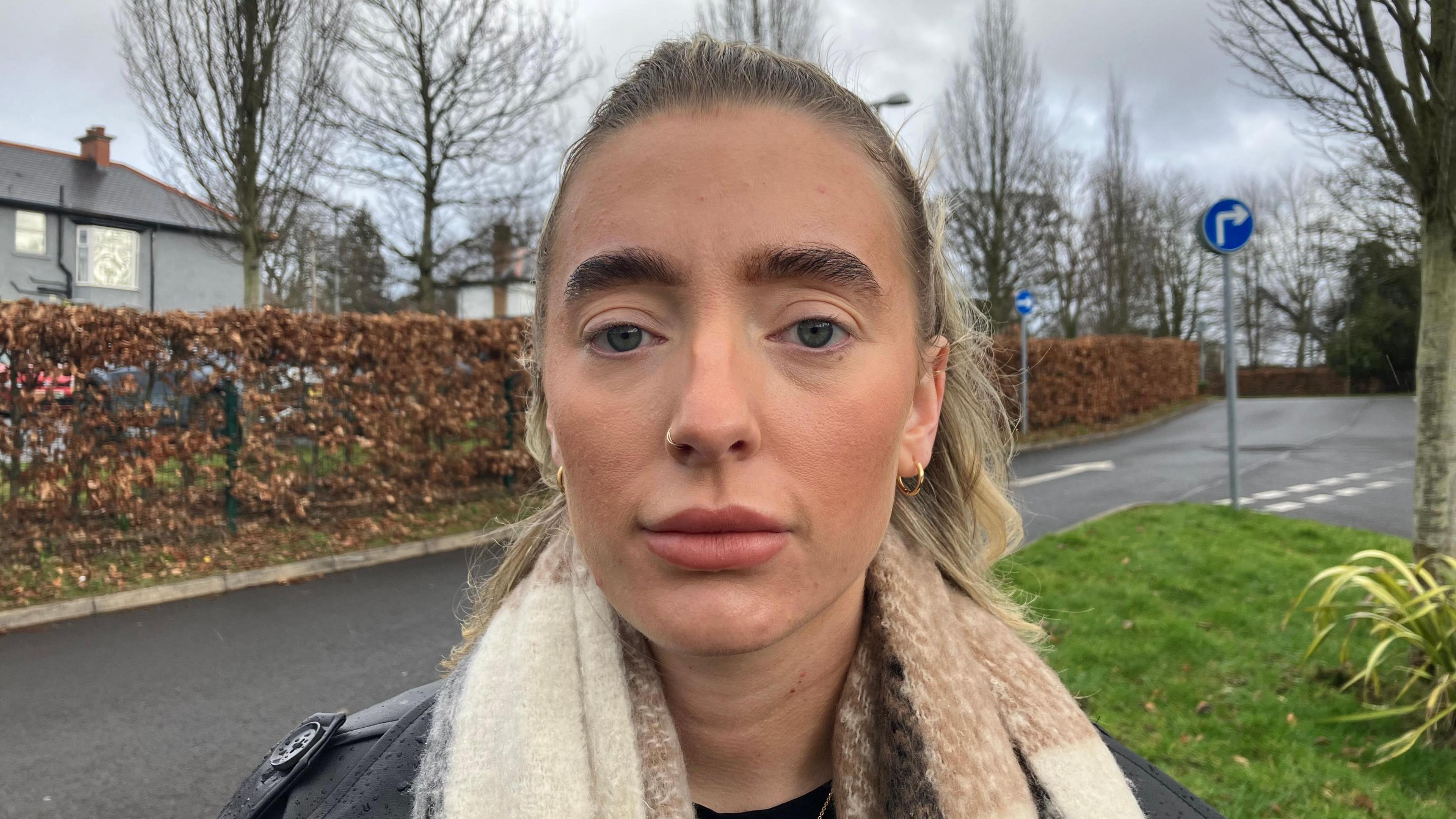 A woman with fair hair that is tied back, off her face. She is staring into the camera, has a nose ring, earrings and wearing a cheque scarf and dark jacket.