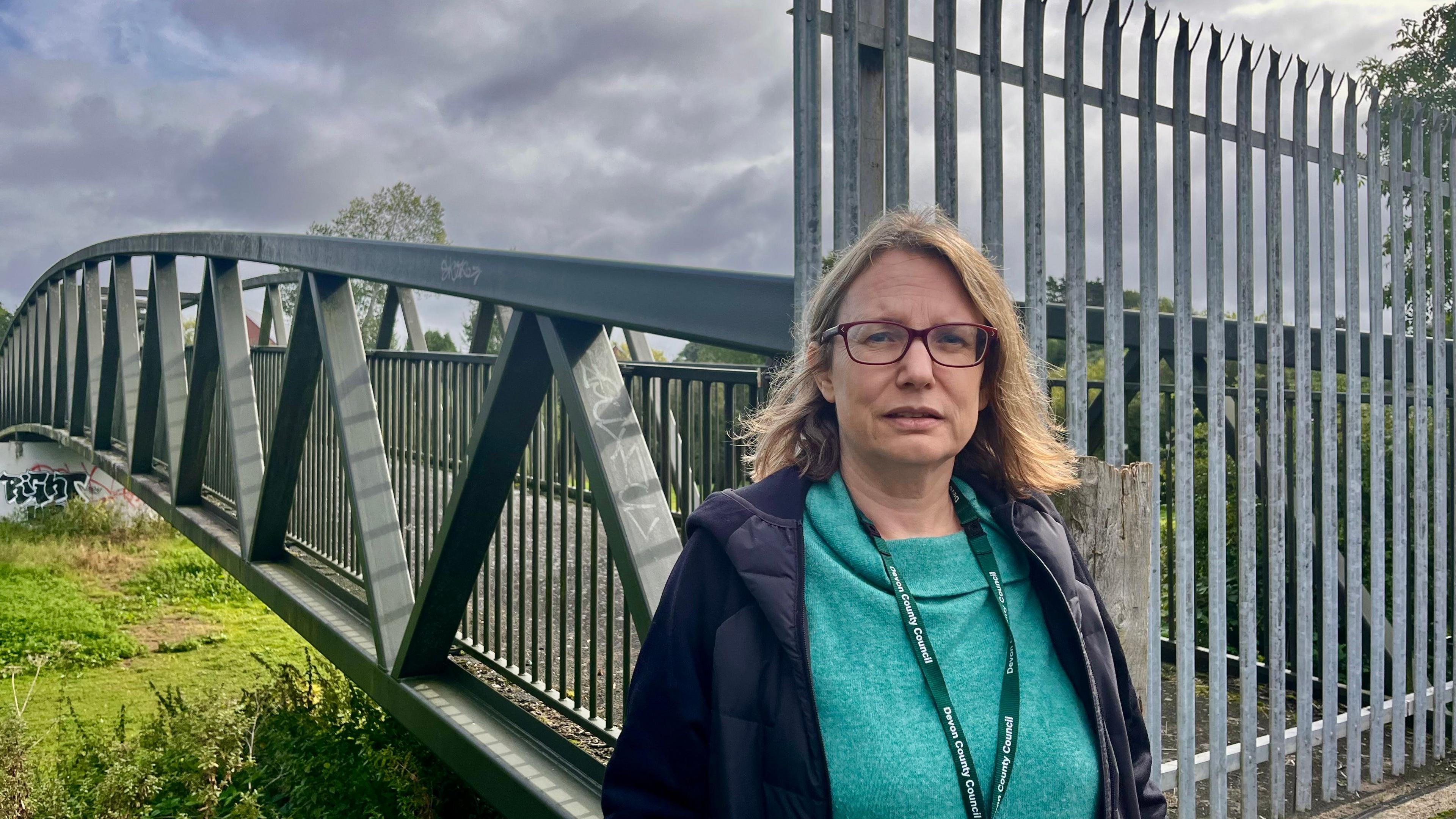 Councillor Jess Bailey standing in front of the closed footbridge, which has high metal fencing at its entrance to prevent people crossing