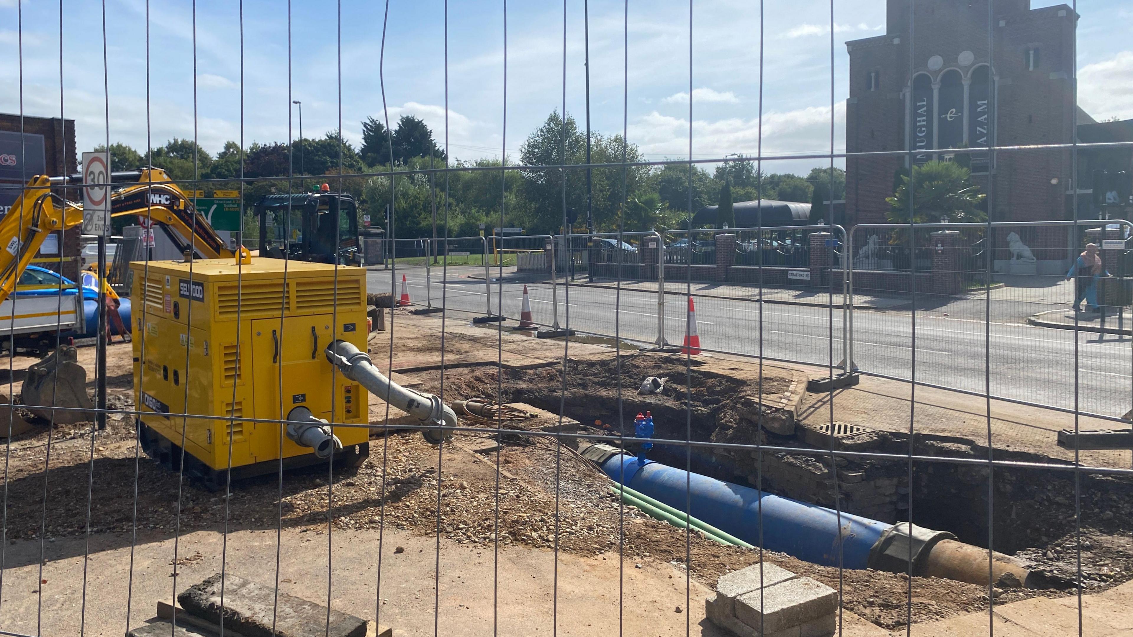 A picture taken behind a fence of a site where engineers are replacing a water main that burst. You can see the pavement has been dug up and there is a visible blue pipe. 