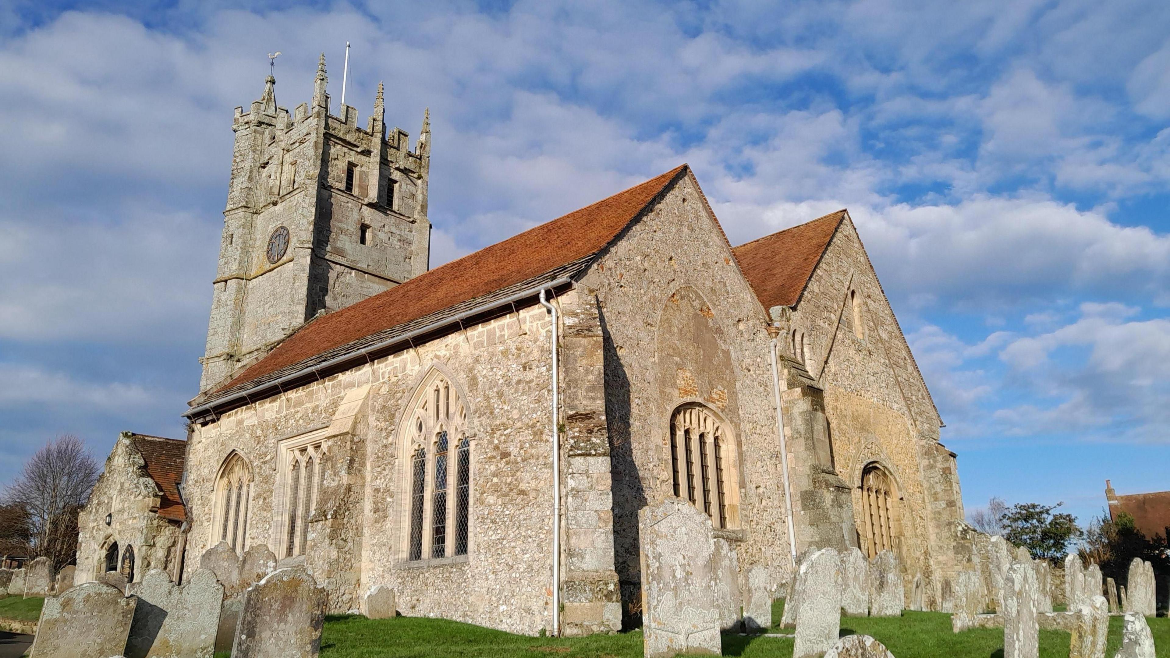 A stone church photographed against a blue sky with white clouds. There are several grave stones in the foreground. The church has an orange tile roof and a bell tower with a flag pole. 