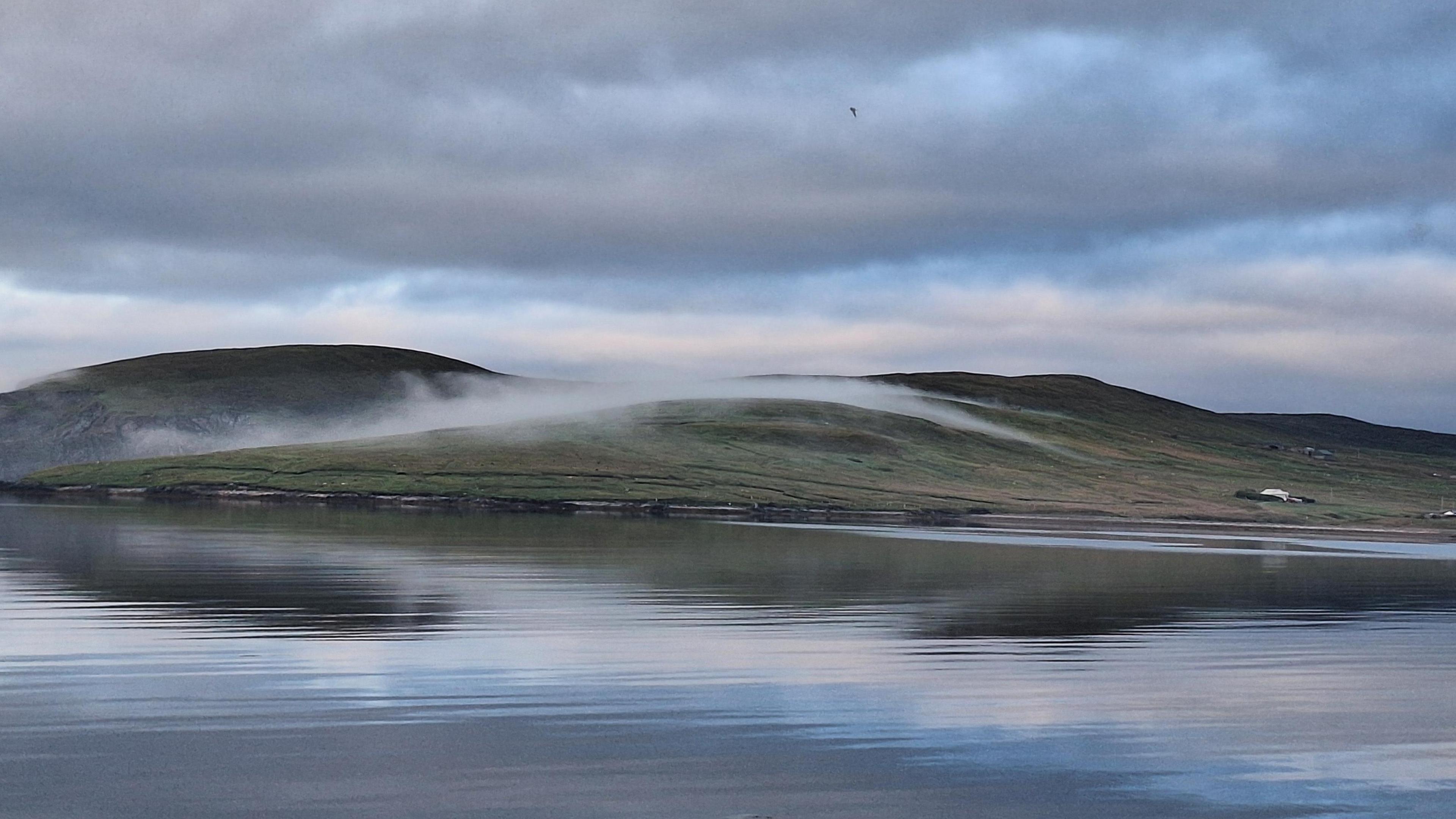 A low hill covered with green grass has low lying grey clouds rolling over the top. There is a calm and still body of water in the foreground of the picture which reflects the blue and grey sky.