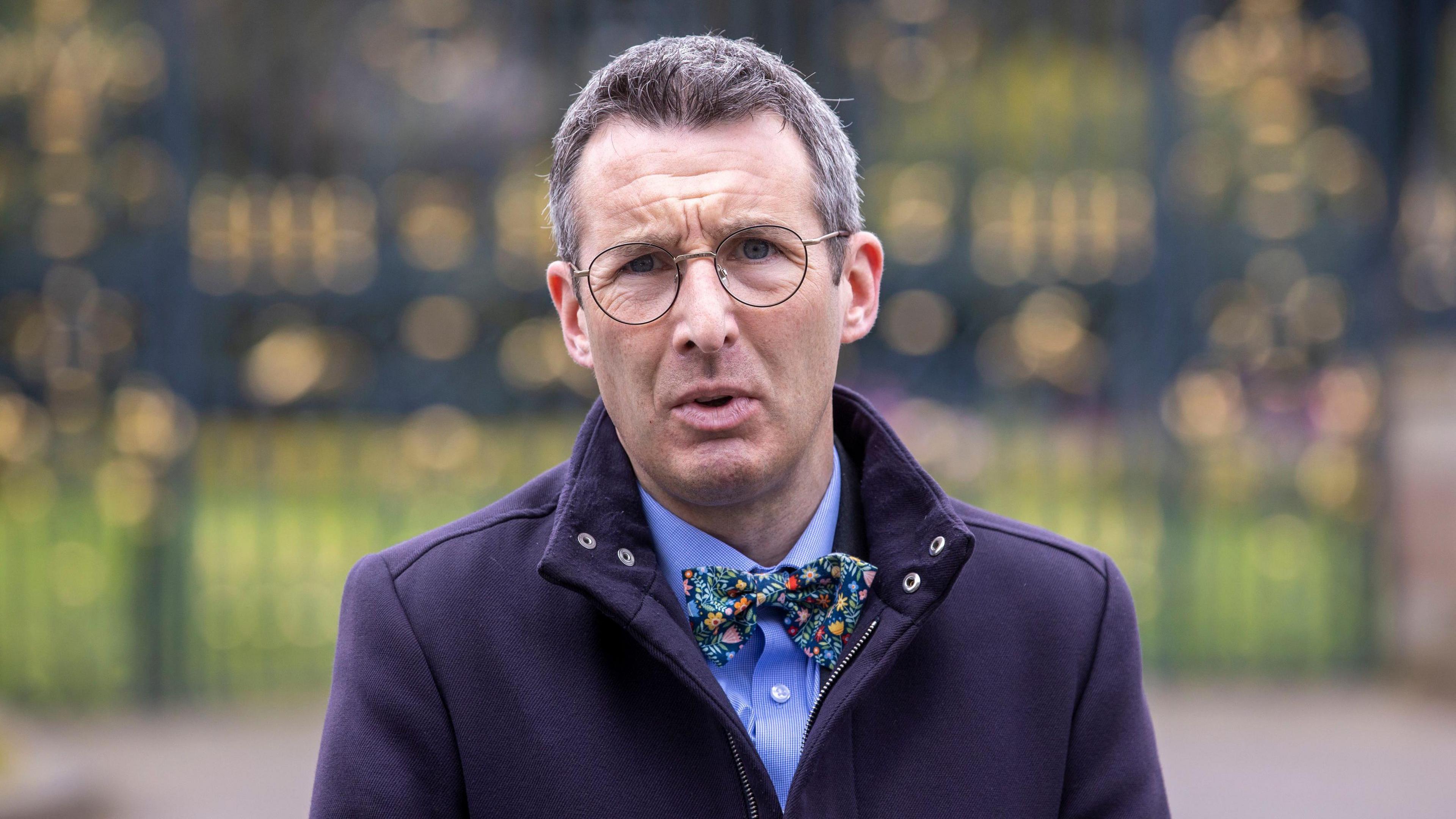 Andrew Muir looking at the camera. He is pictured from the chest up. He has grey hair and is wearing a blue shirt, a bow tie, navy jacket and round glasses. He has a serious expression on his face. The background of the image is blurred. 