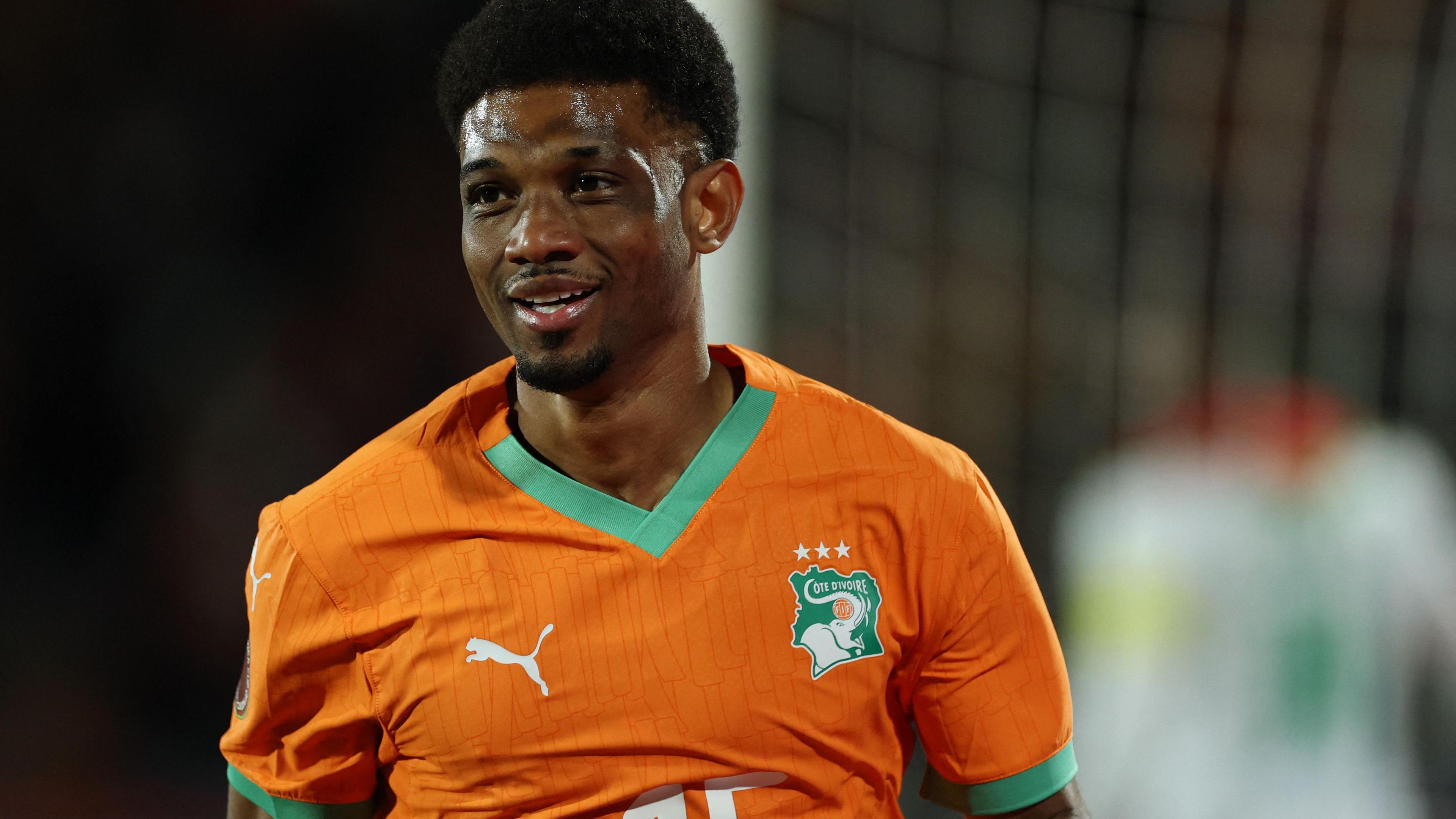 Amad Diallo, seen from the chest up wearing an orange Ivory Coast shirt with green trim, breaks a slight smile on his face. Blurred in the background behind him can be seen part of a goalpost, a net and the back of a Burkina Faso player in white